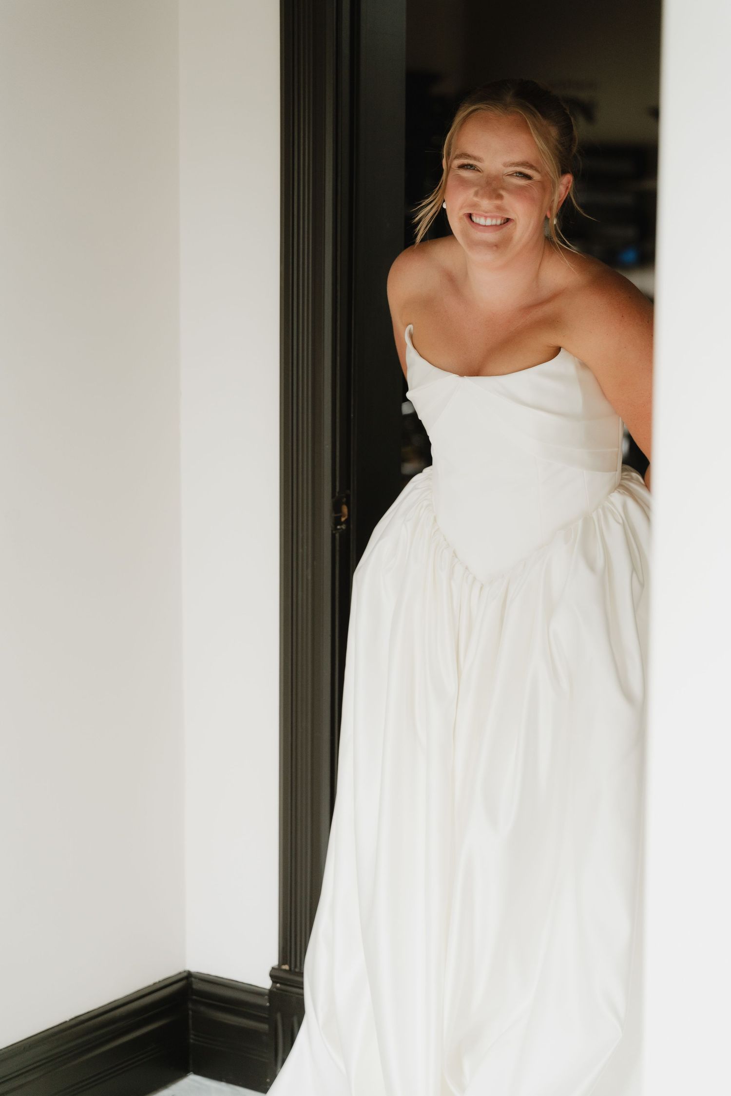 Smiling bride in a strapless white wedding gown peeks through a doorway with black trim.