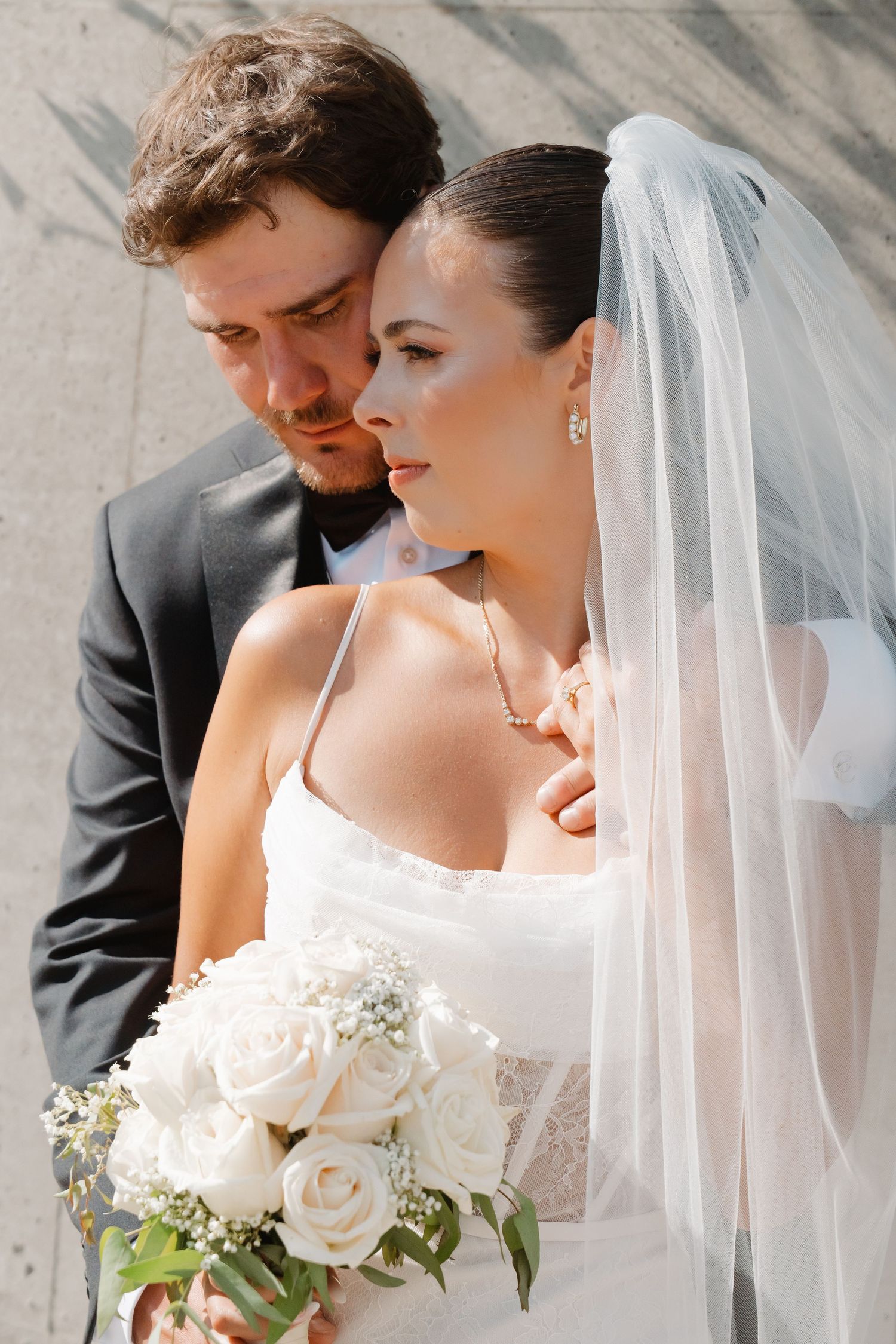Bride in white gown and veil holds rose bouquet as groom in black suit embraces her tenderly outdoors.