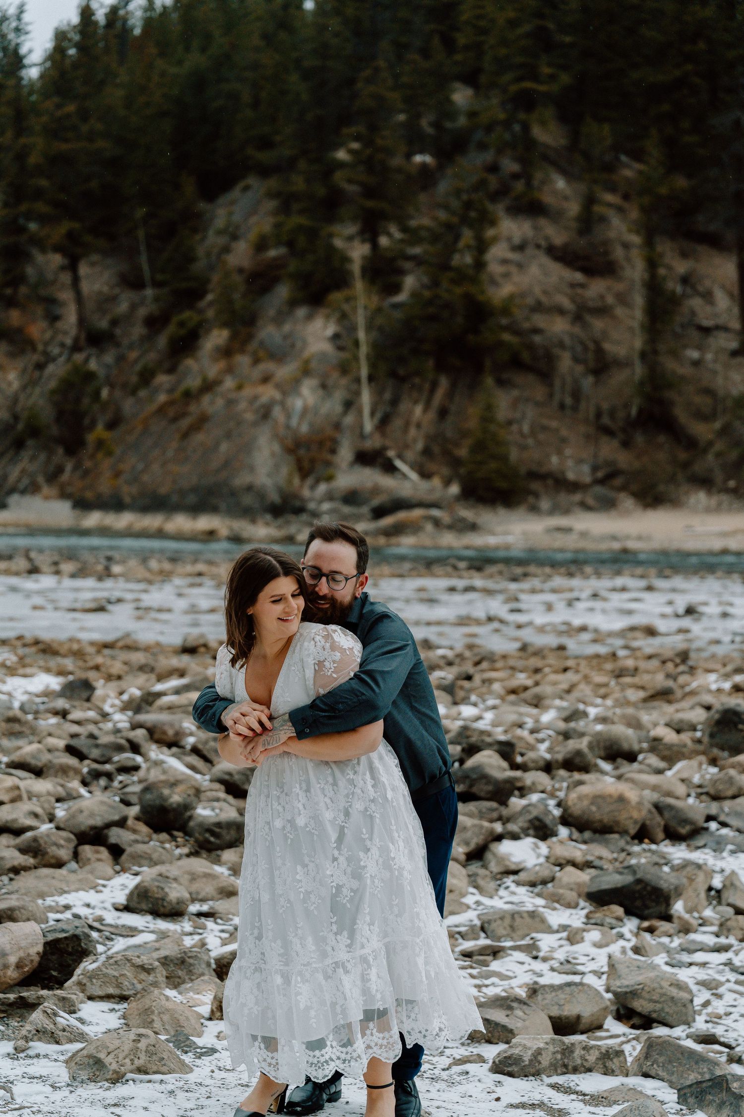 Intimate wedding portraits on a rocky beach with evergreen trees and mountains in the background.