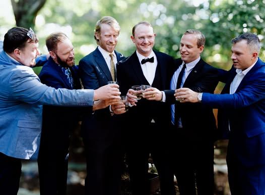 Groomsmen in blue suits and bowties sharing drinks and laughing outdoors.