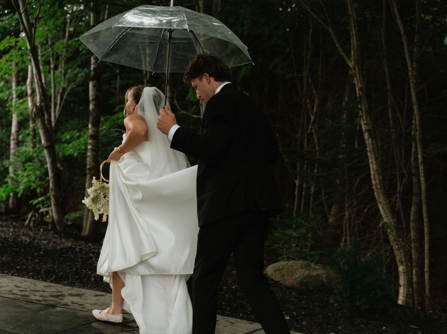 Bride and groom walking in rain under clear umbrella on their wedding day, surrounded by lush green trees.