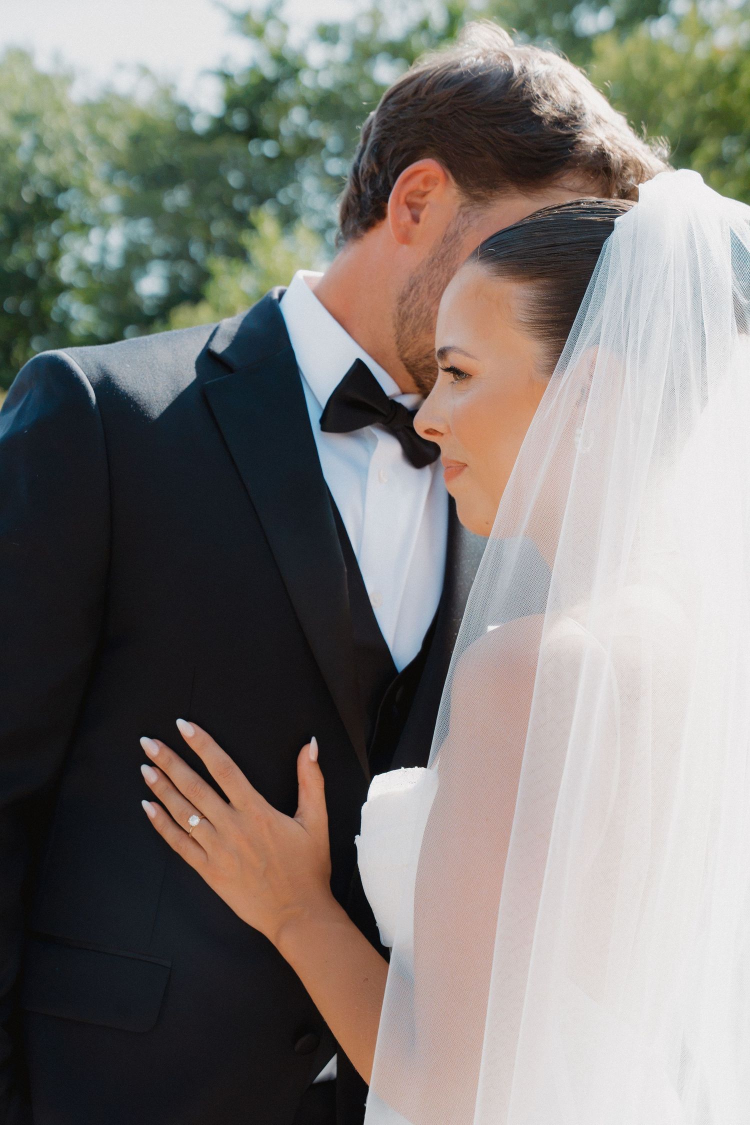 Bride in white veil rests hand on groom's chest in black tuxedo, sharing intimate moment outdoors.
