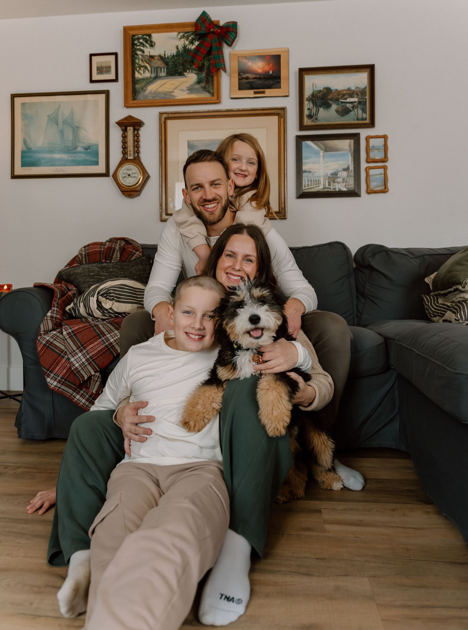 Family relaxing on gray couch with their fluffy white and brown dog in living room with framed artwork.