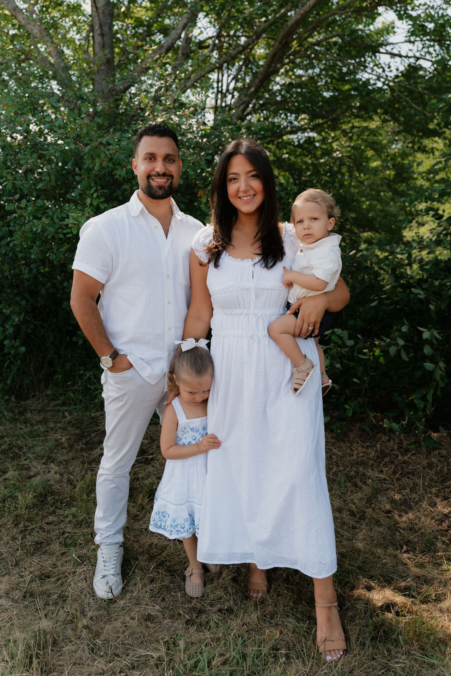Happy family of four in matching white outfits posing outdoors with lush green trees in the background.