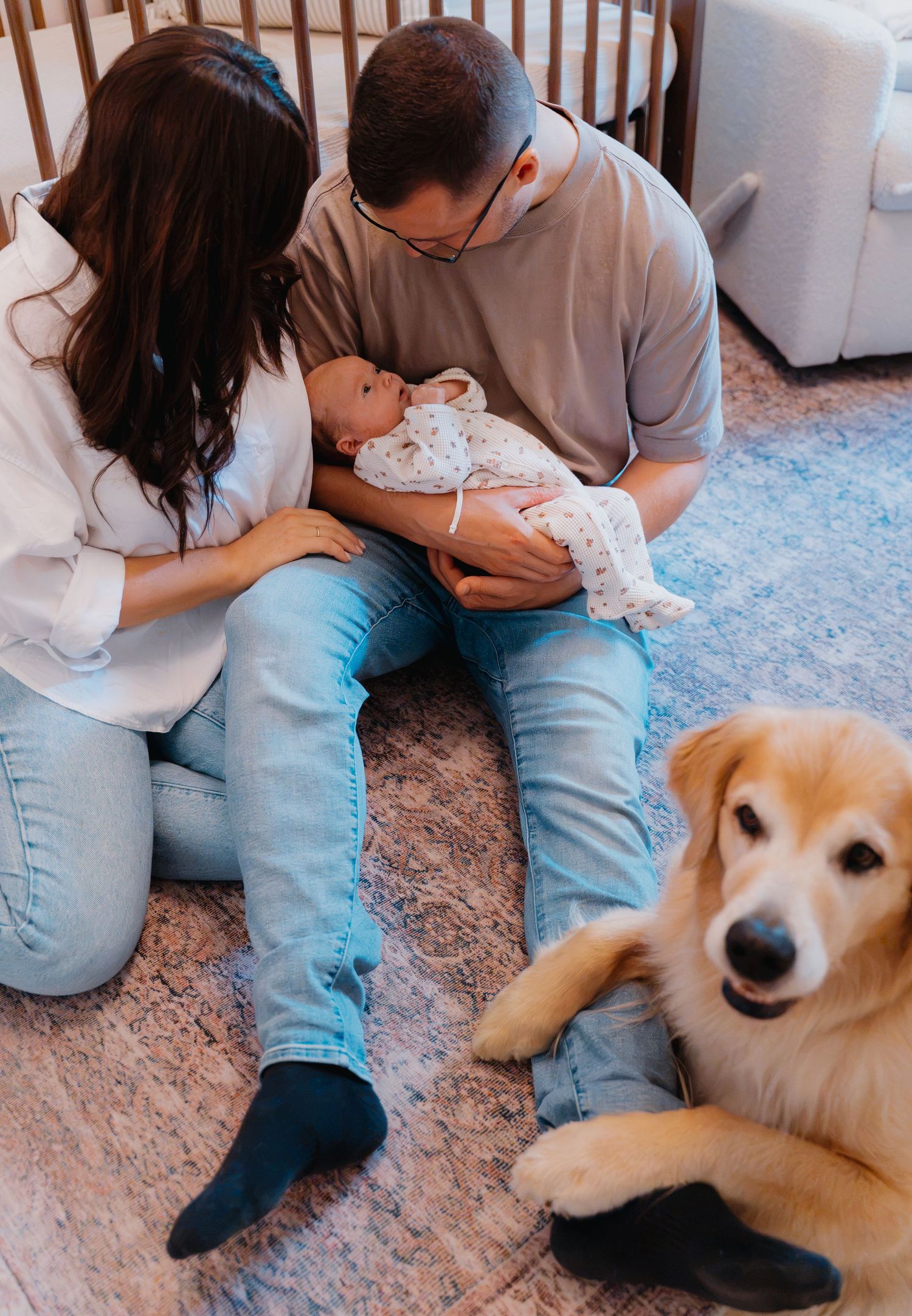 New parents sit on floor holding newborn baby with golden retriever dog beside them in cozy nursery room.