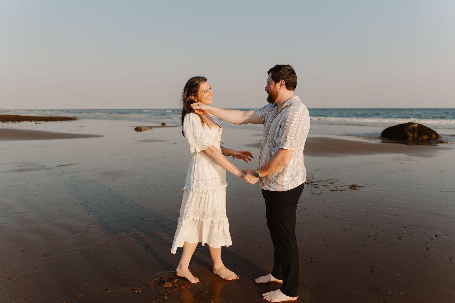Couple dancing romantically on a sandy beach at sunset, woman in white dress, man in casual shirt and black pants.