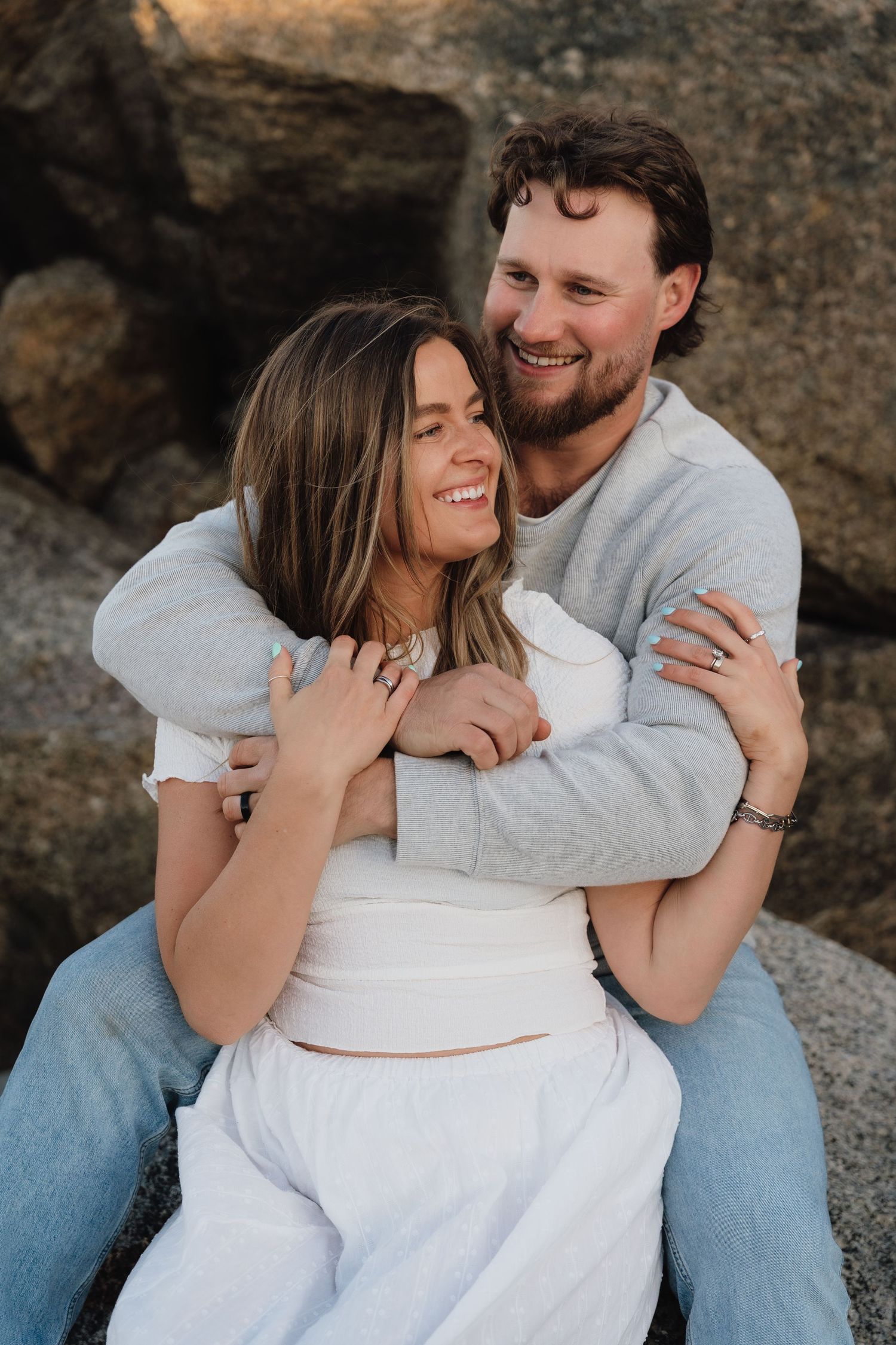 Couple embracing and laughing on rocky shore, man hugging woman from behind, both smiling joyfully outdoors.
