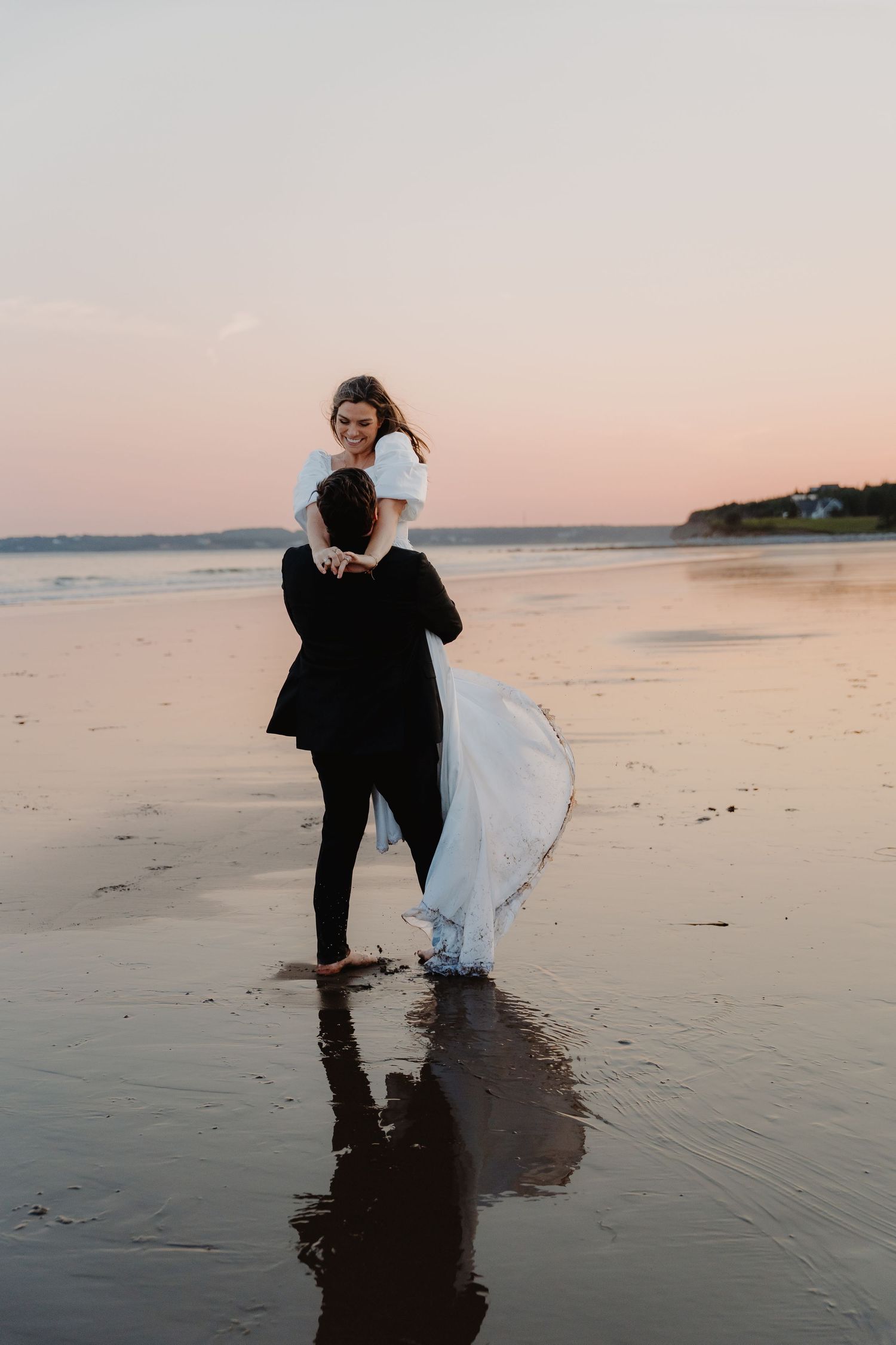 Groom lifting bride in white gown on a reflective beach at sunset, waves and pink sky in the background.