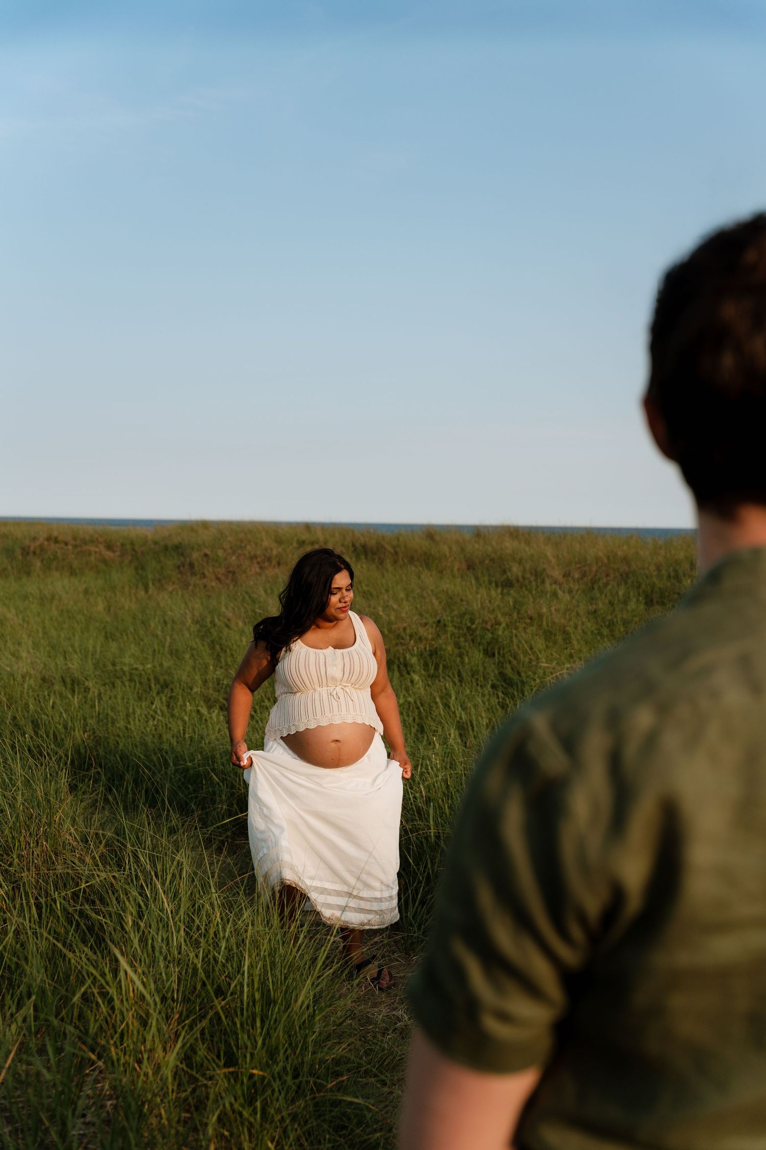 Pregnant woman in white dress walks through green field toward man in olive shirt during golden hour outdoor maternity shoot.
