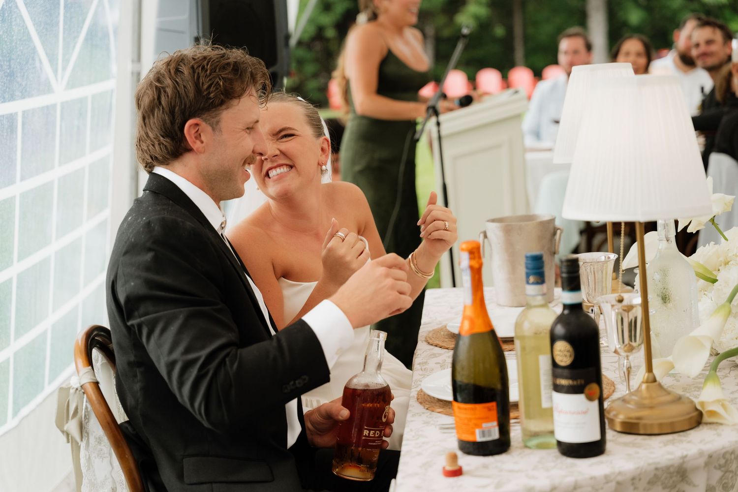 A laughing bride and groom sit at a wedding reception table with champagne bottles and glasses during toasts.