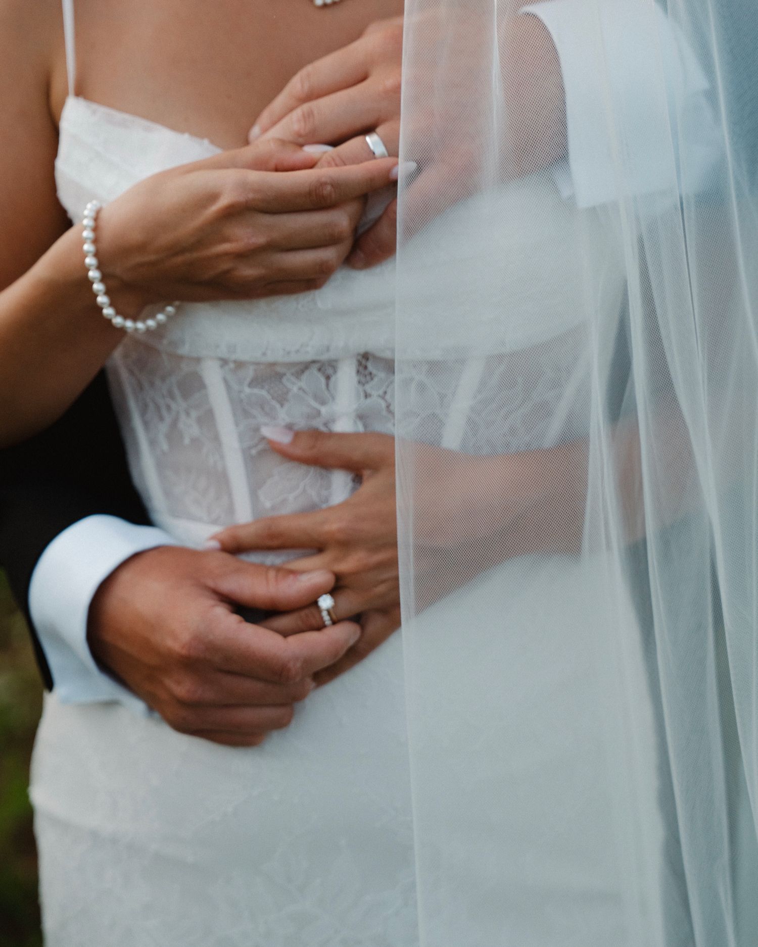 Close-up of bride and groom holding hands, showcasing wedding rings, pearl bracelet, and lace wedding dress with flowing veil.