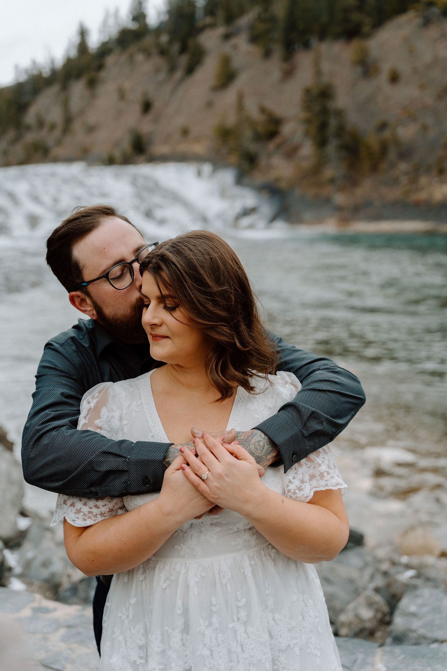 Couple embraces during romantic mountain lakeside photo session in wedding attire.