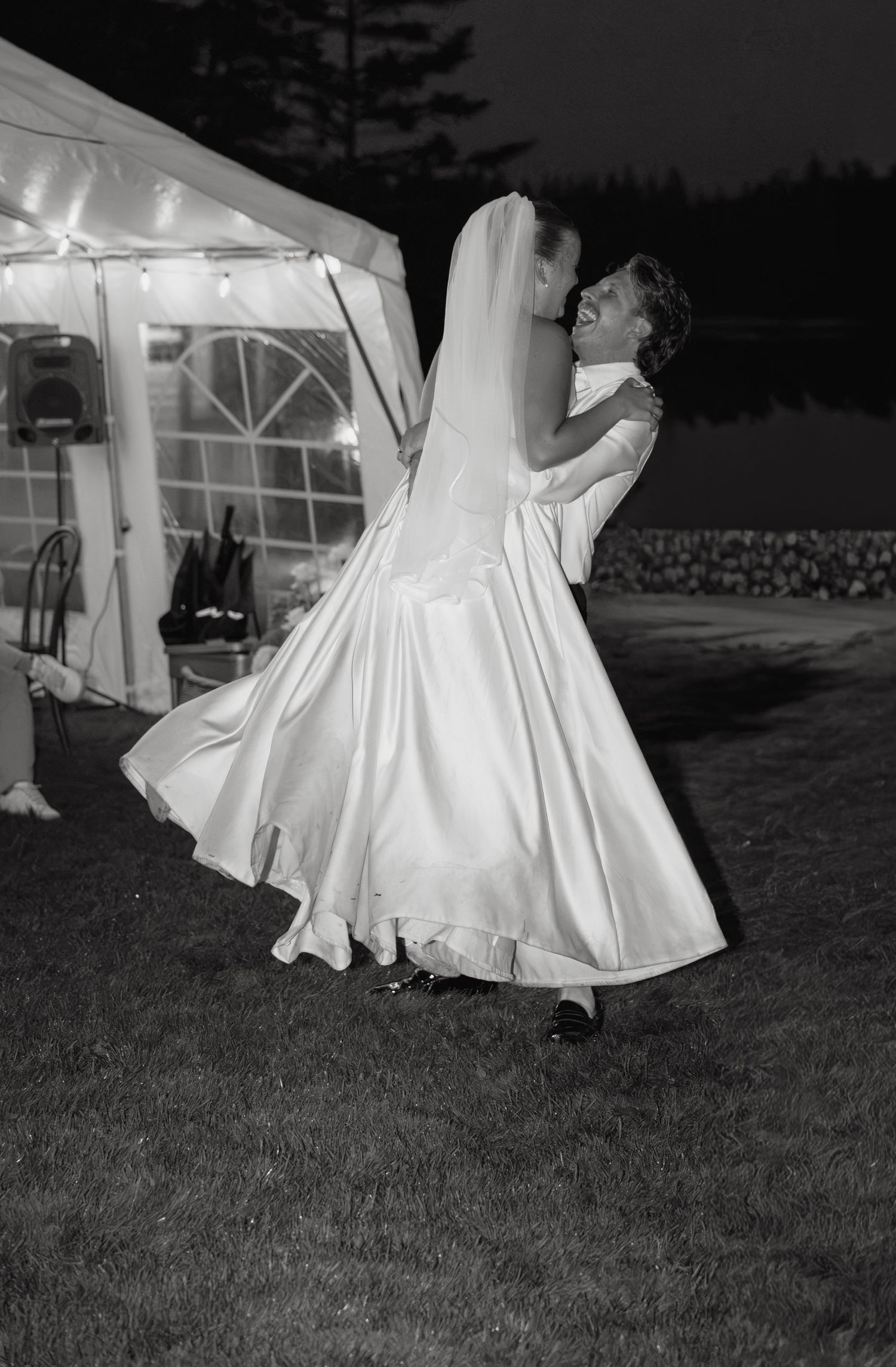 Black and white photo of groom lifting bride in flowing white gown outside wedding reception tent.
