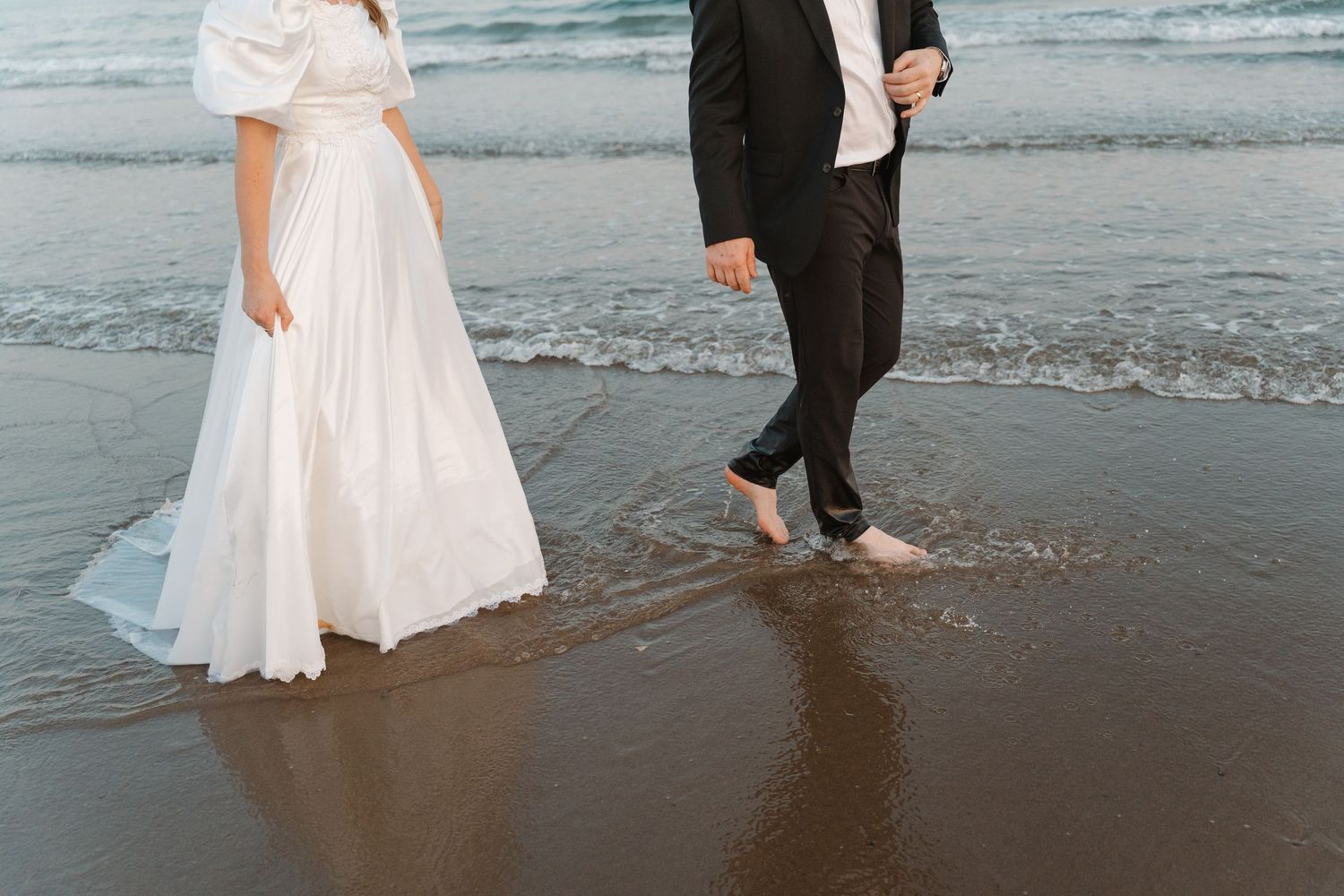 Bride in white gown and groom in black suit walking barefoot on a sandy beach with ocean waves.