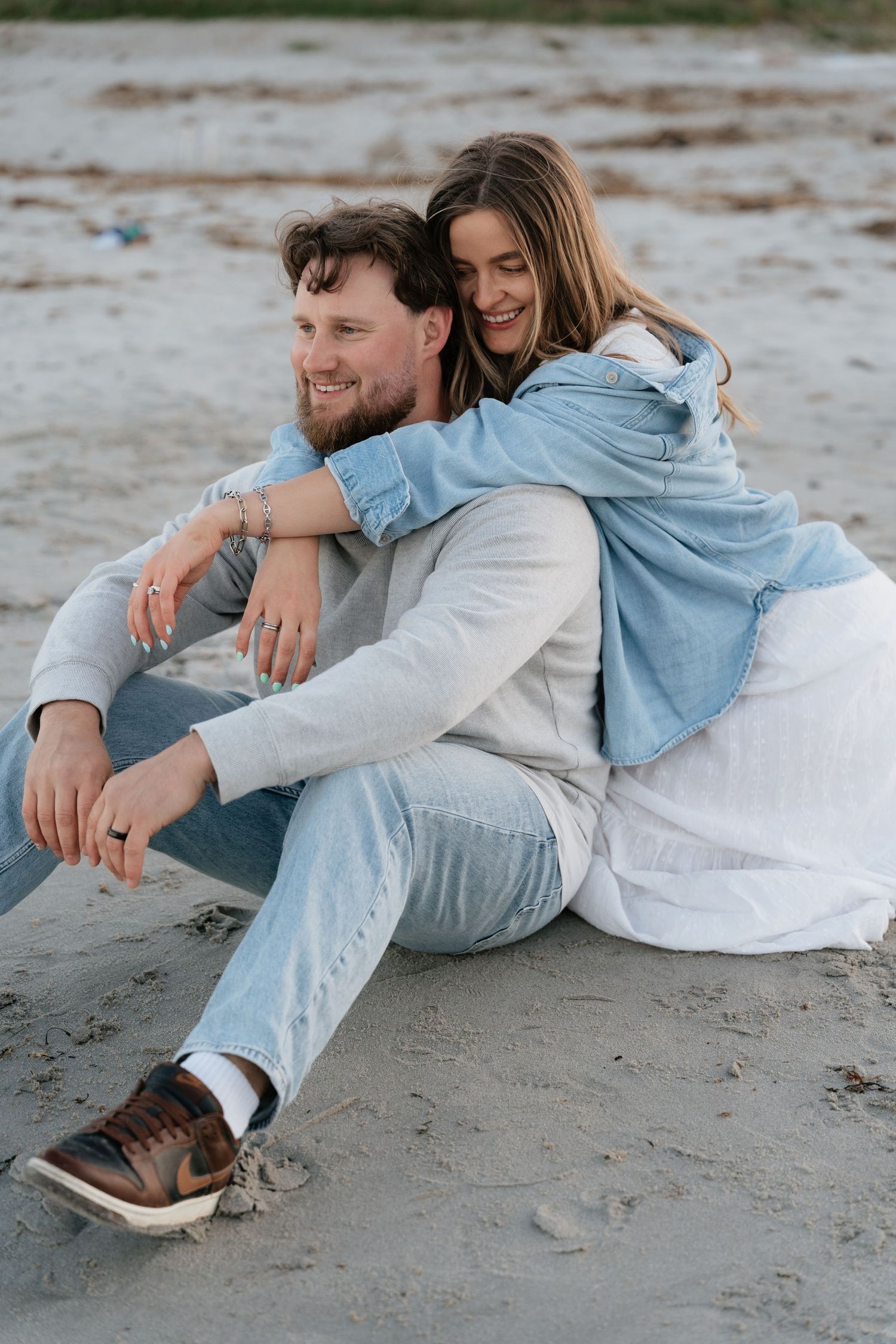 Happy couple sitting on sandy beach, woman hugging man from behind, both smiling, wearing casual denim outfits.