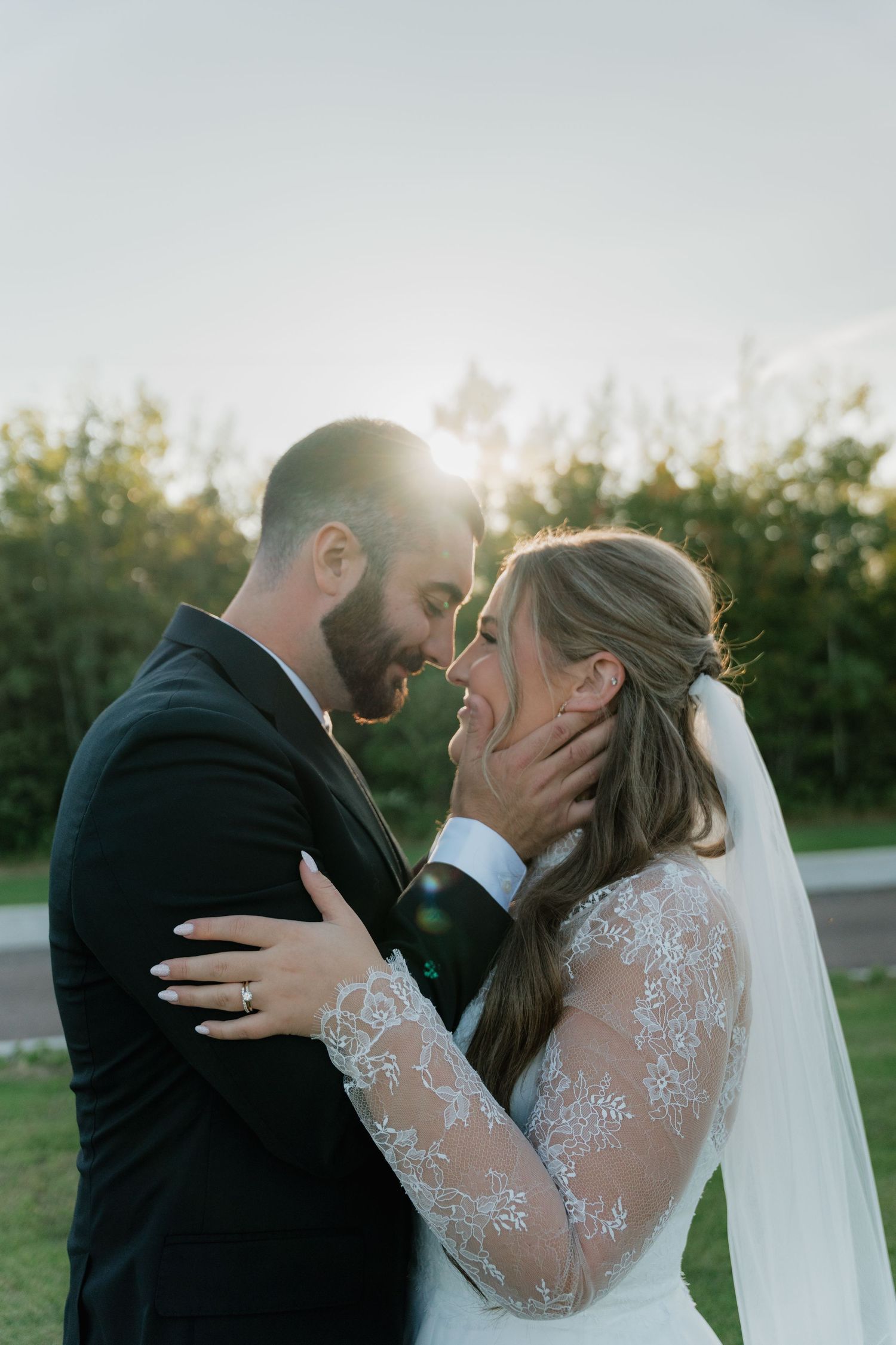Bride and groom share a romantic forehead touch outdoors, glowing in golden sunset light, bride in lace dress with veil.