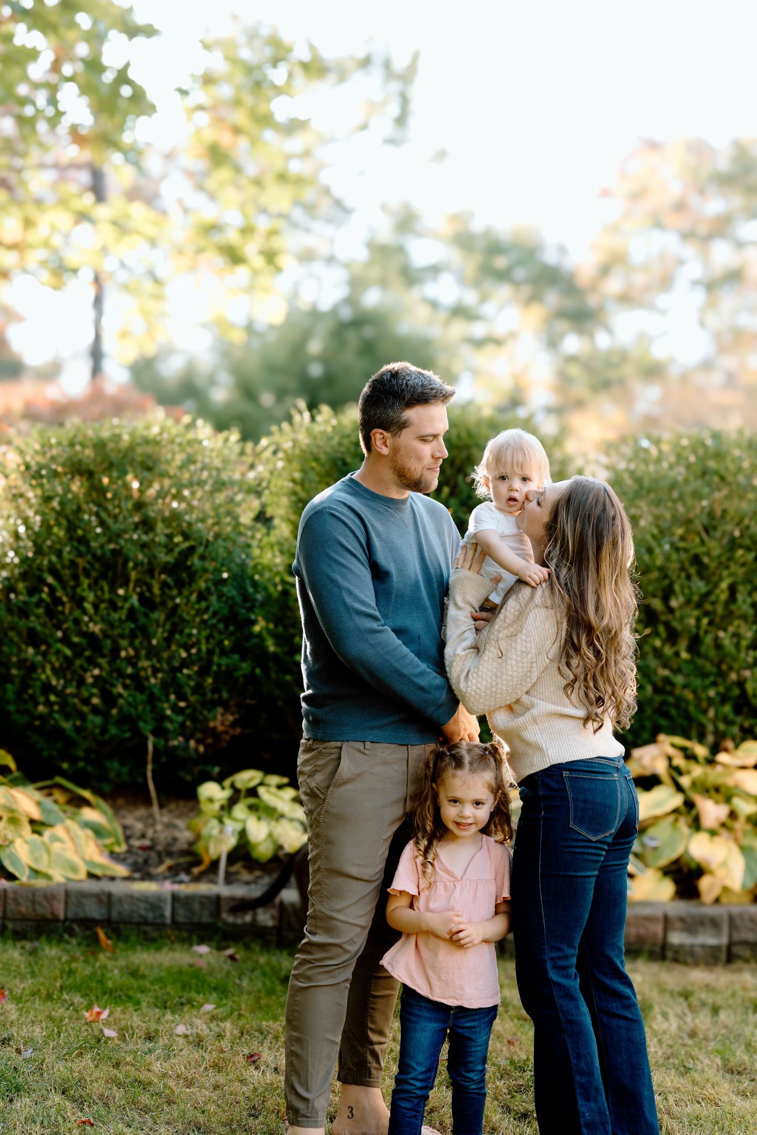 Family shares tender moments together during an outdoor autumn photoshoot in front of green hedges.