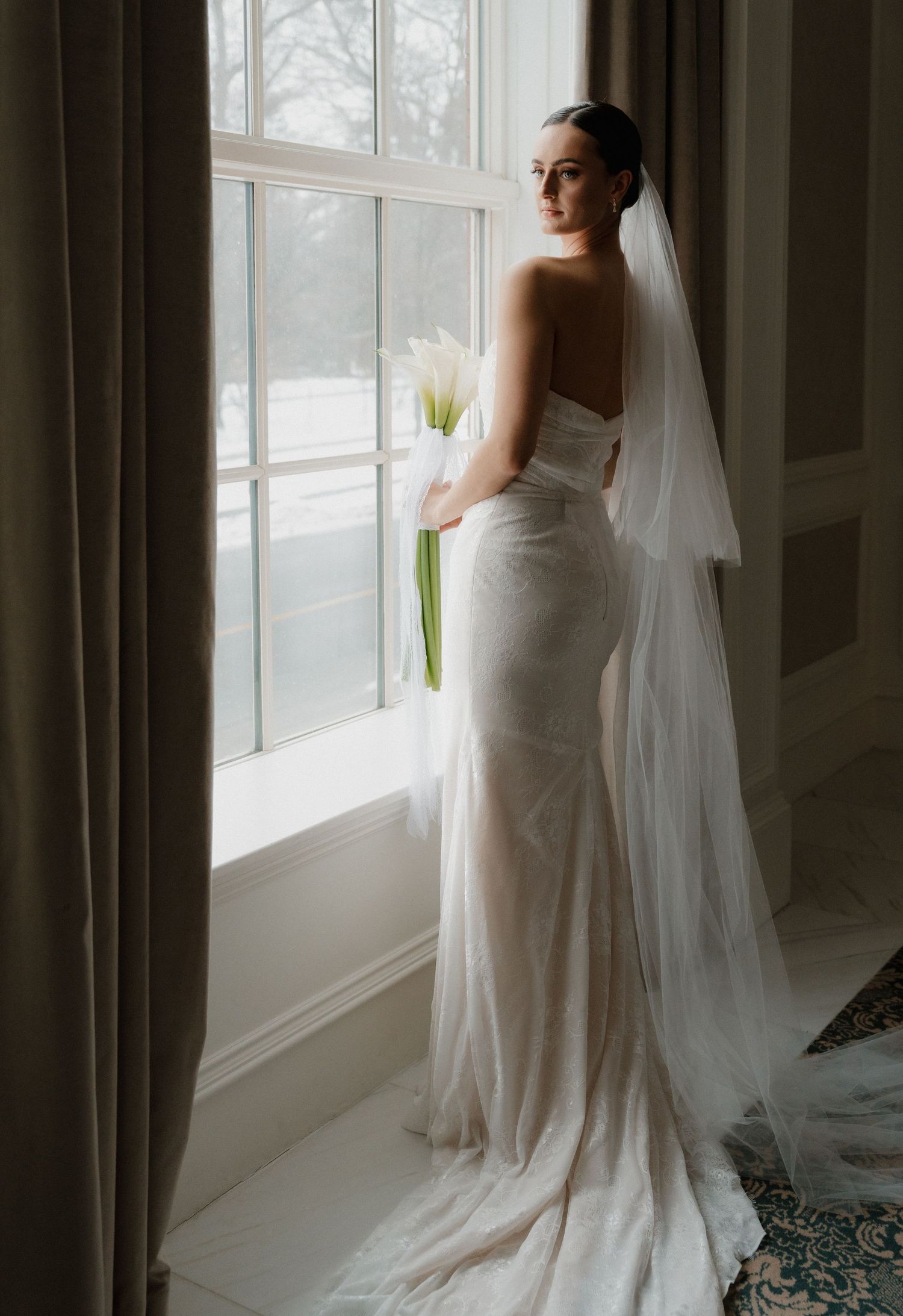 Bride in elegant white wedding dress and veil stands by large windows in dramatic natural lighting.