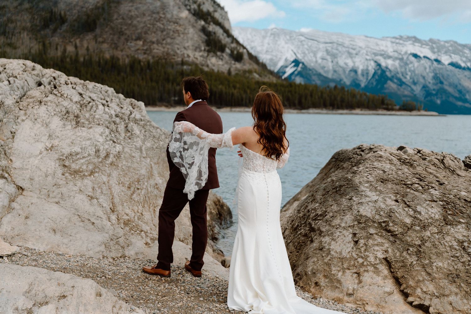 A couple embraces on a rocky mountain overlook with snow-capped peaks and a lake in the distance.
