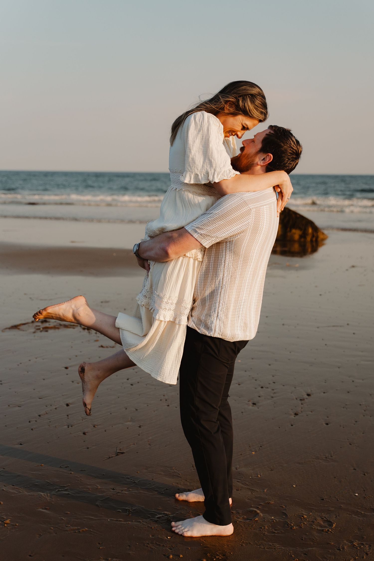 A couple shares a romantic embrace on a sandy beach at sunset, with the man lifting the woman in white.