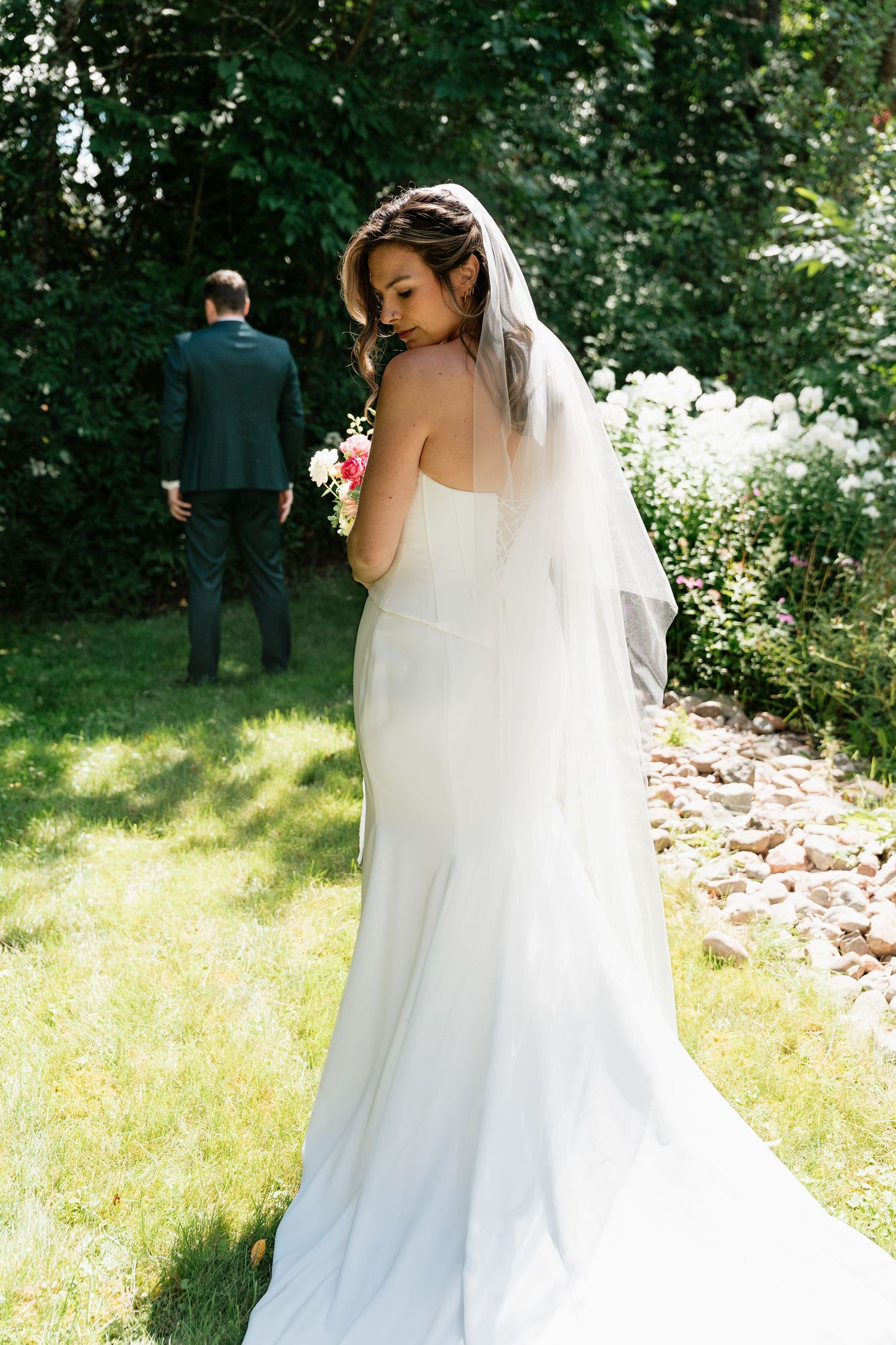 Bride in white gown and veil holds bouquet outdoors, groom in dark suit stands nearby among lush green trees.