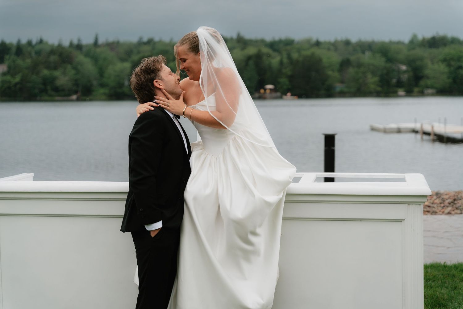 Bride and groom sharing a romantic kiss near a lakeside venue, bride in white gown with veil, groom in black tuxedo.