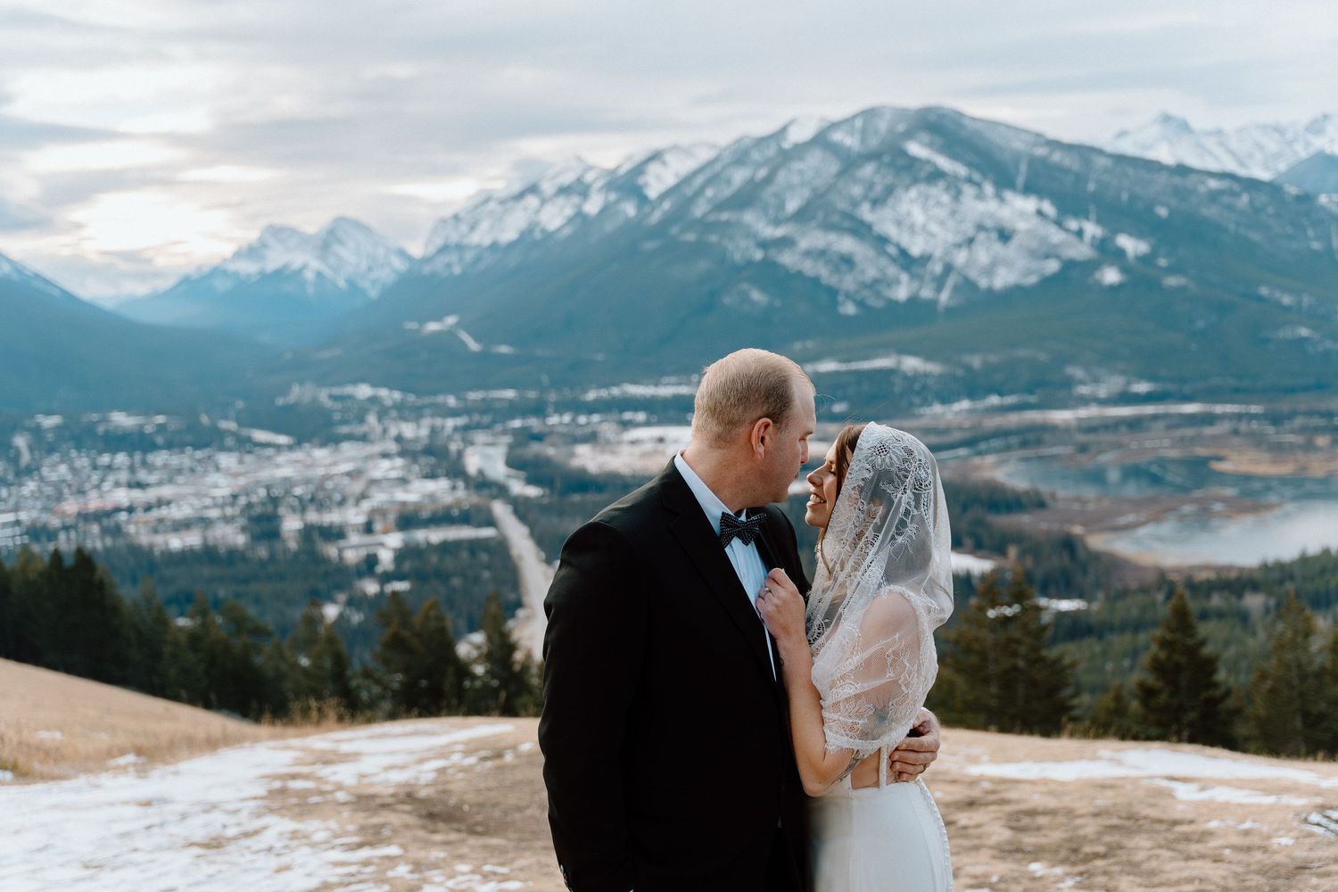 A couple shares a romantic kiss on a mountain overlook with snowy peaks and a scenic winter valley in the background.