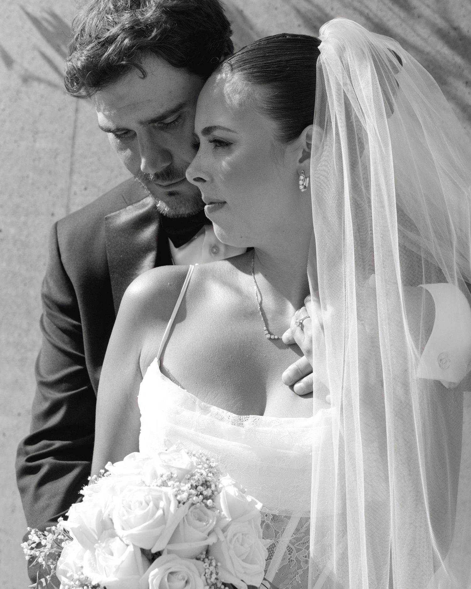 Black and white wedding photo of bride in veil holding roses bouquet with groom in suit embracing her.