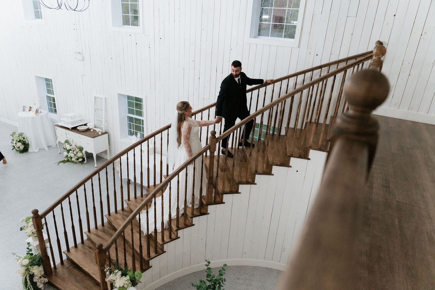 Bride and groom descend elegant wooden staircase in white barn wedding venue adorned with floral arrangements.