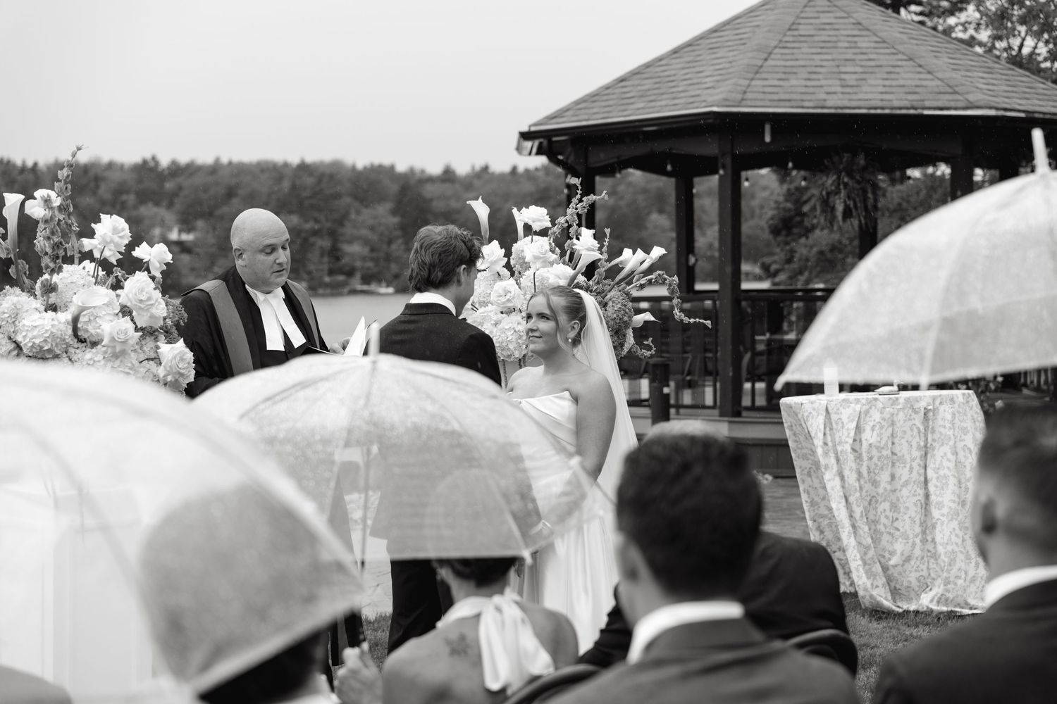 Outdoor wedding ceremony in the rain with guests holding umbrellas, bride and officiant at altar near gazebo.