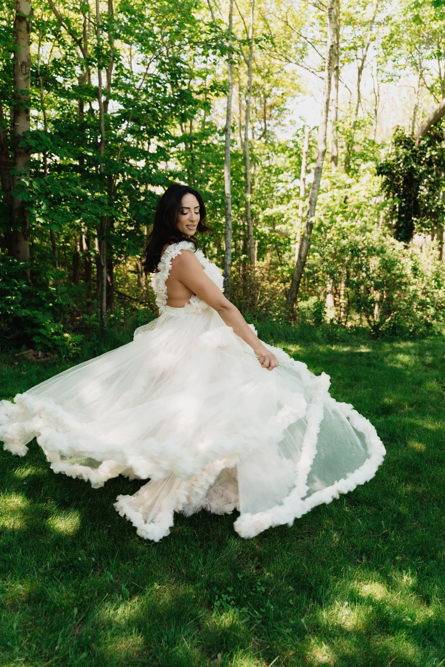 Bride twirling in a flowing white ballgown with ruffled trim in a lush green forest setting.
