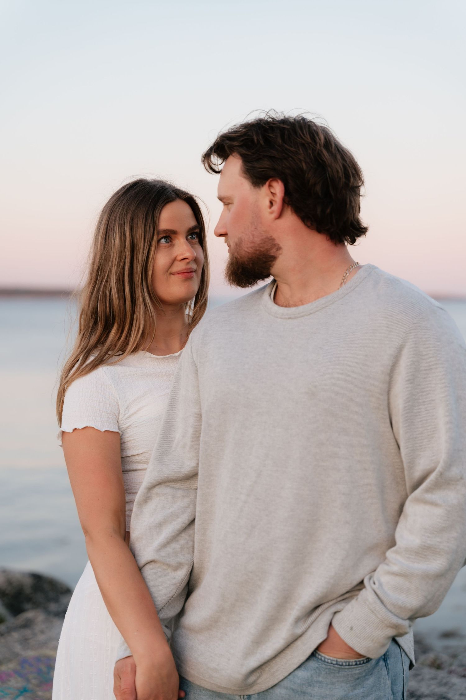 Couple sharing a romantic moment on a rocky beach at sunset, woman in white dress, man in gray sweater.