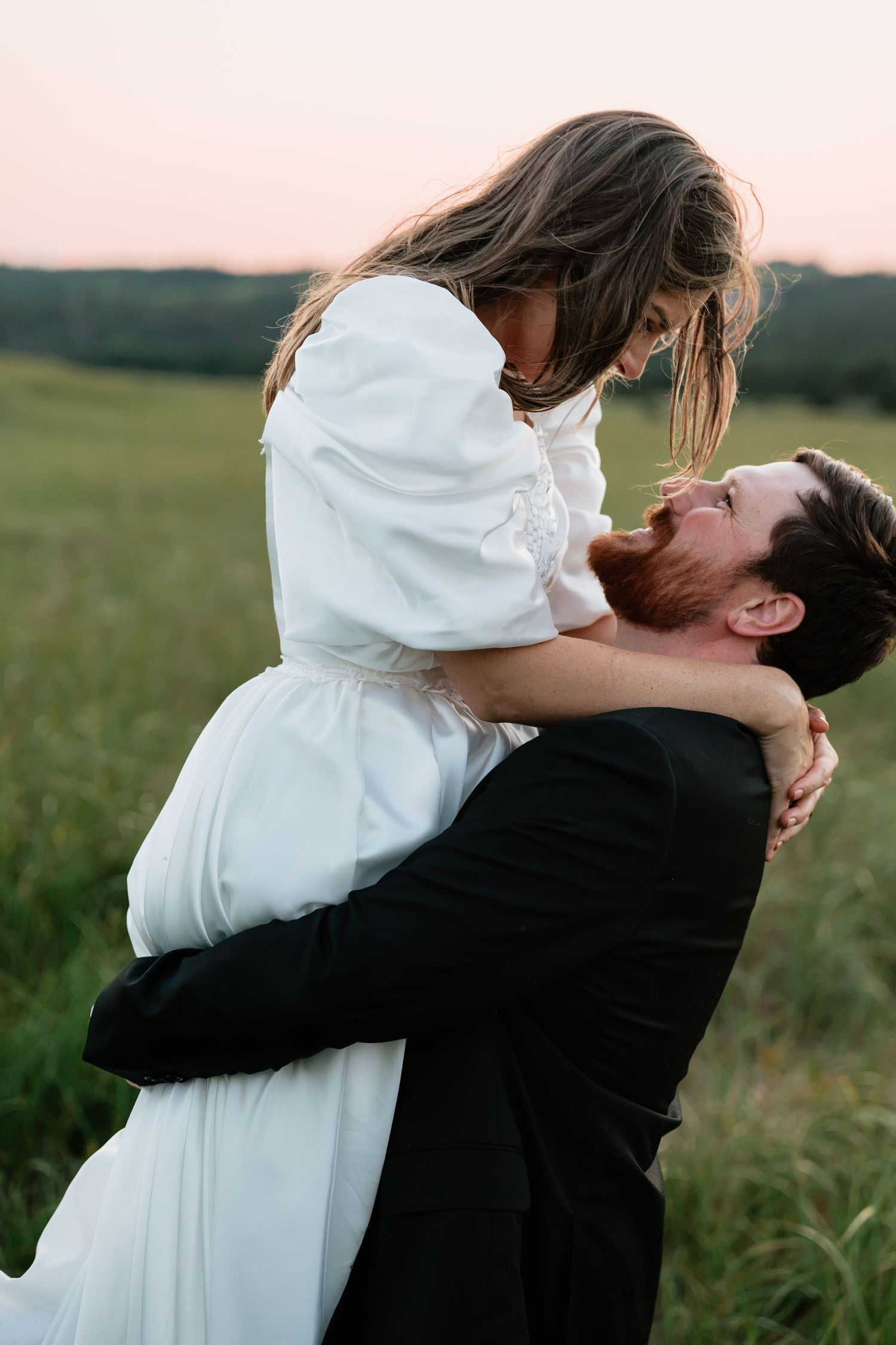 Groom lifting bride in white dress in a green field at sunset, sharing a romantic moment.
