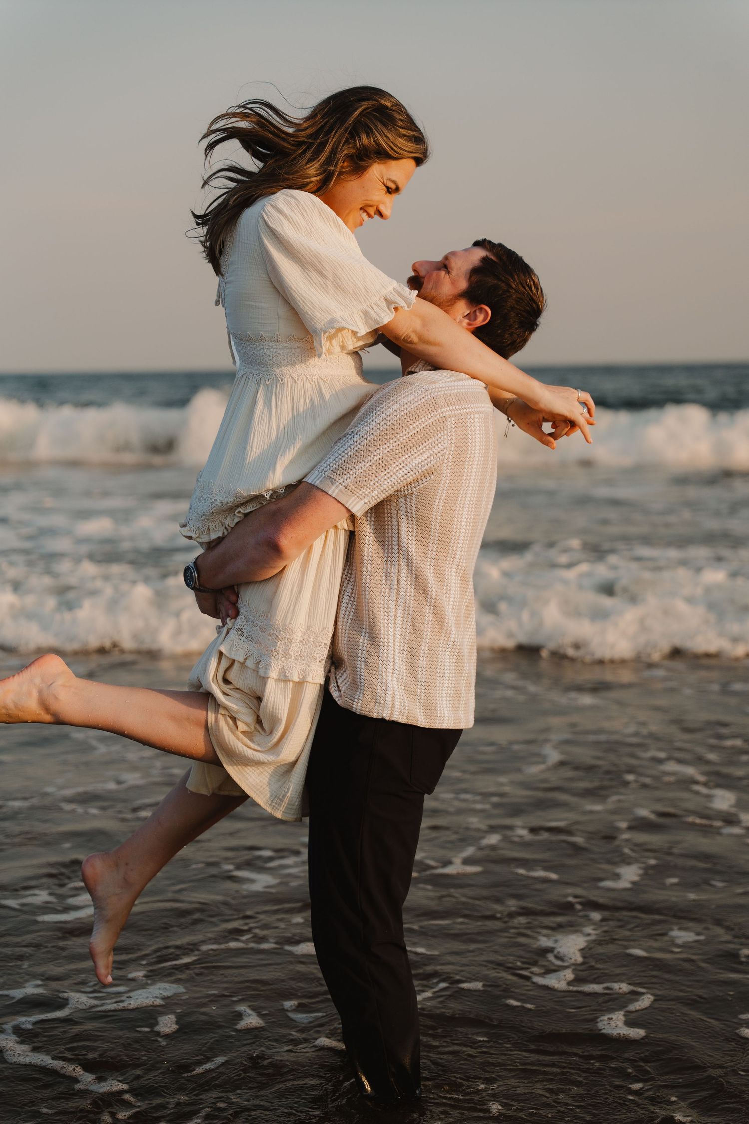 A couple shares a romantic moment on the beach at sunset, as the man lifts the woman in a white dress by the waves.