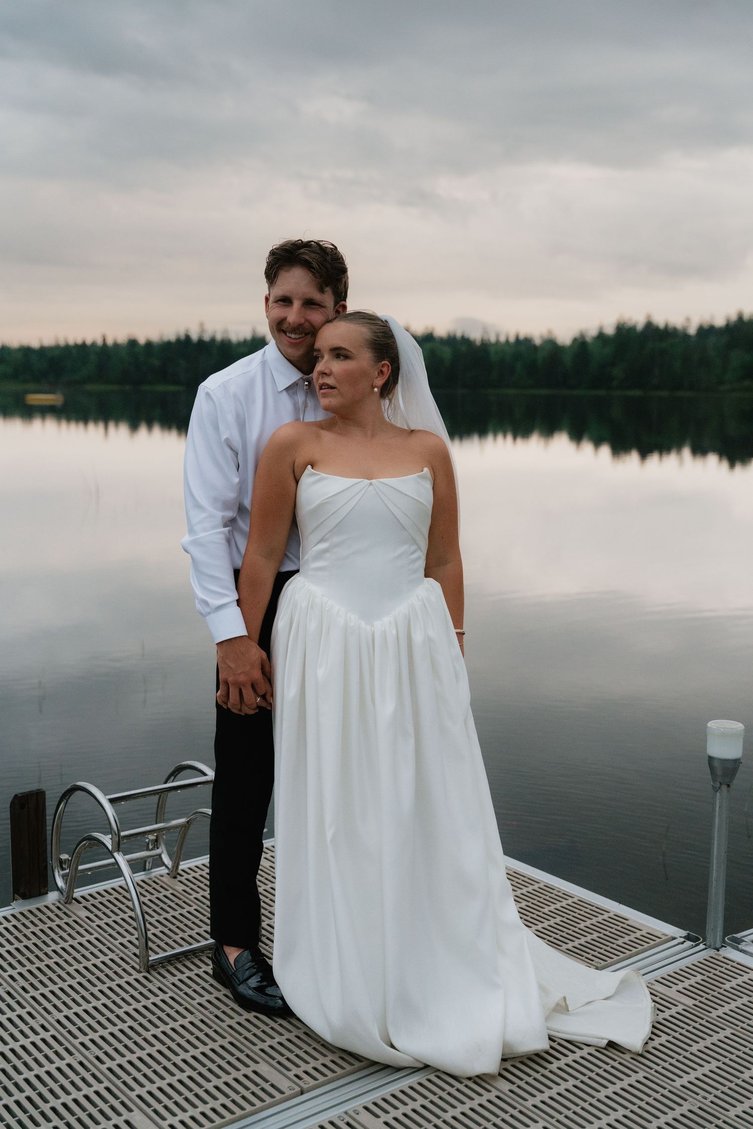Bride and groom holding hands on a lakeside dock at sunset with calm water and forest reflections.