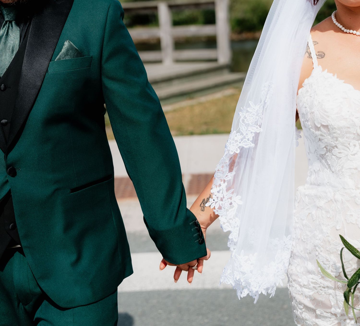 Bride in white lace gown and veil holds hands with groom in dark green suit during outdoor wedding ceremony.