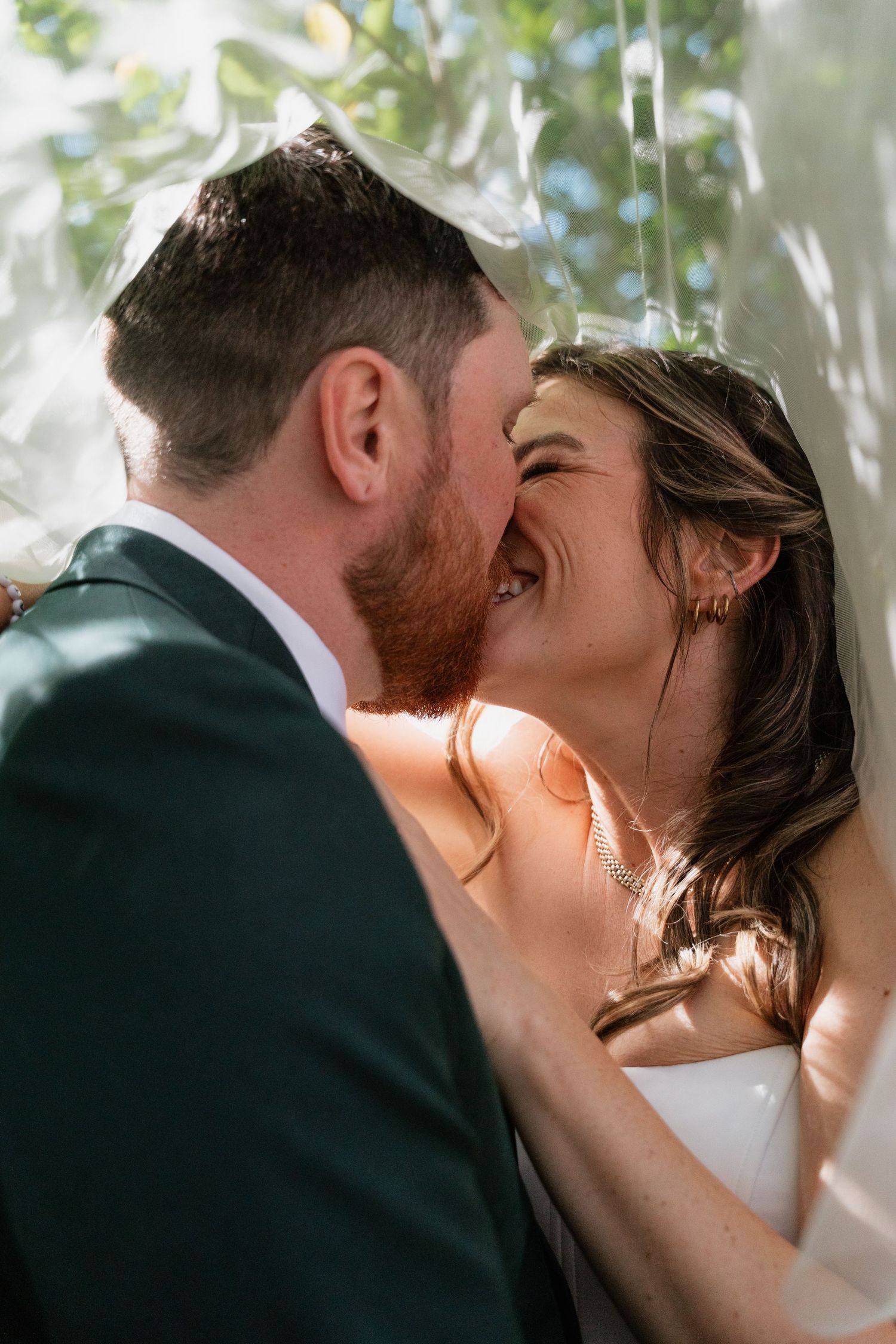 A joyful bride and groom share a laughing kiss under a white veil outdoors on their wedding day.