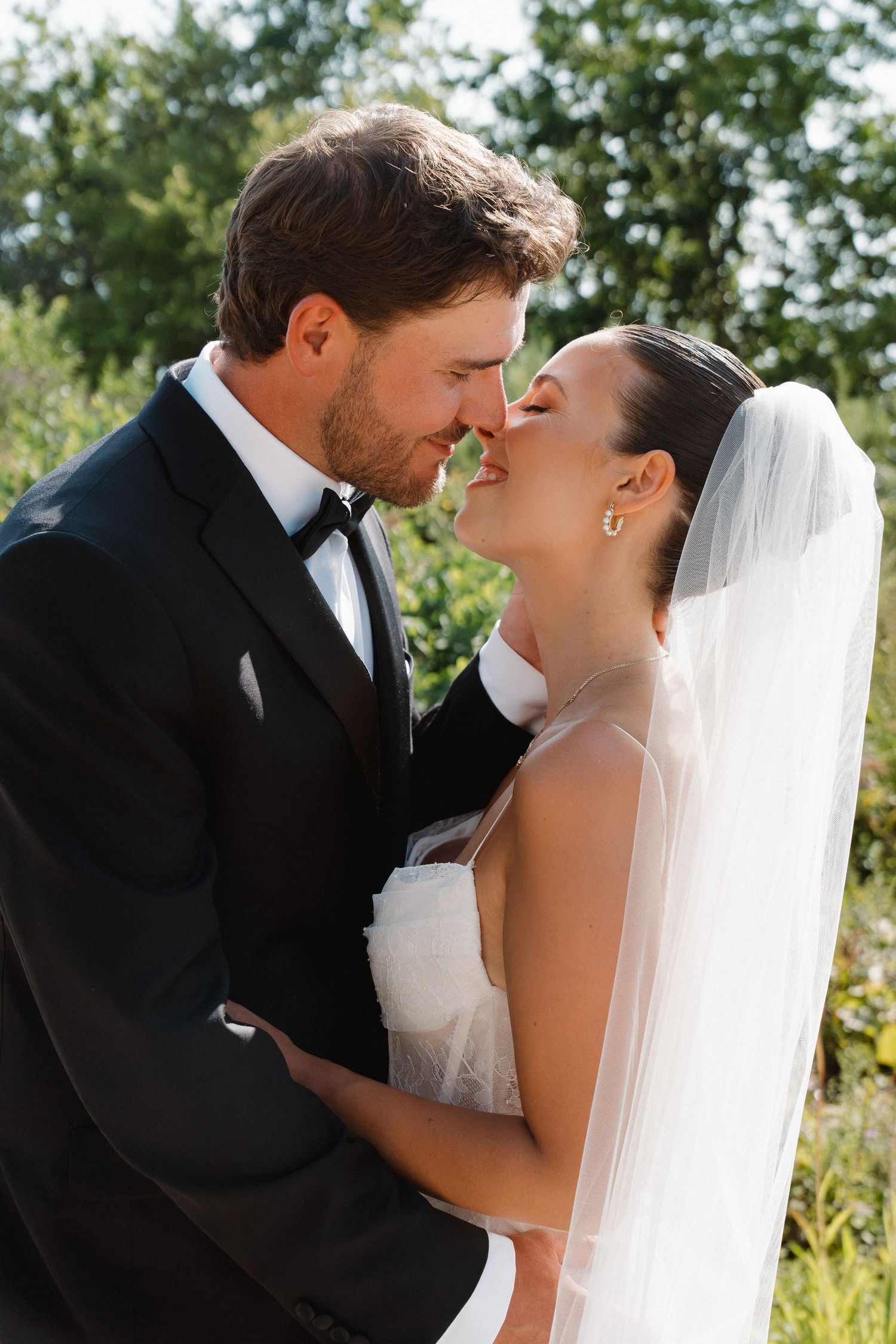 A bride and groom share a romantic moment outdoors, nearly kissing, dressed in a white gown with veil and black tuxedo.