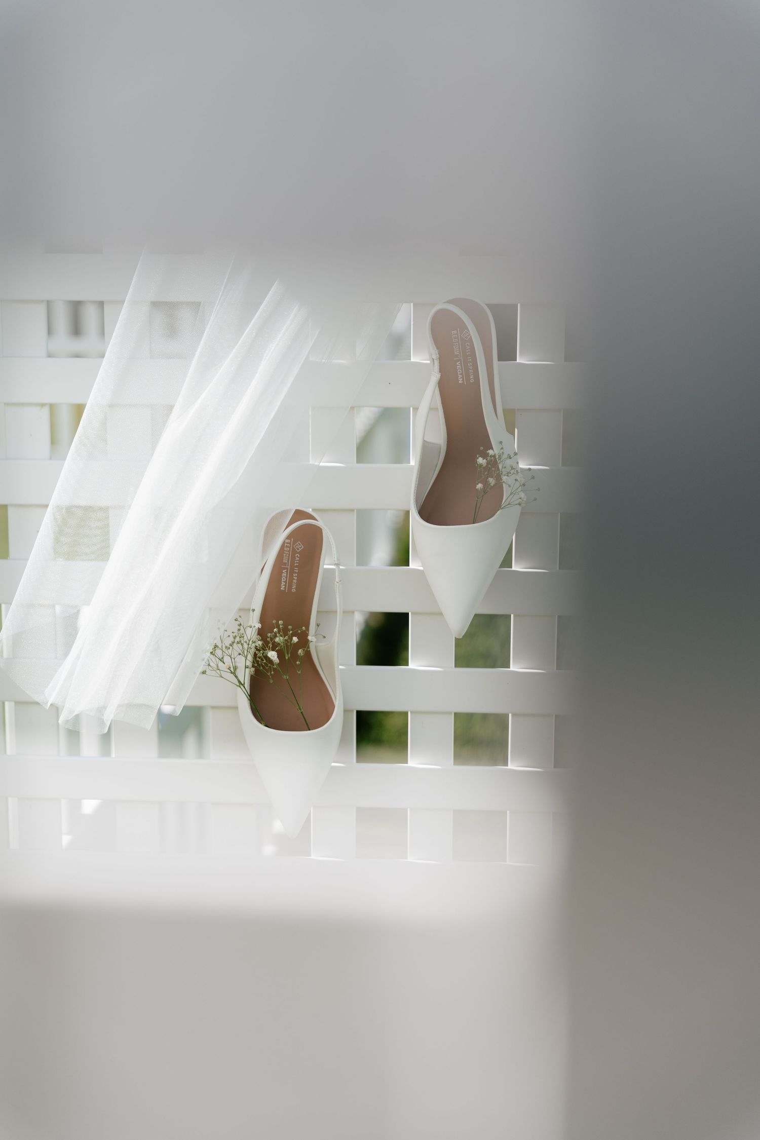 White bridal heels with baby's breath flowers displayed on a lattice fence beside a flowing wedding veil.
