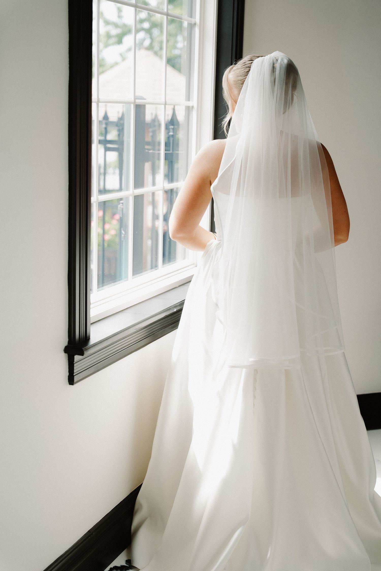 Bride in white strapless gown and veil gazing out a large window with black trim.