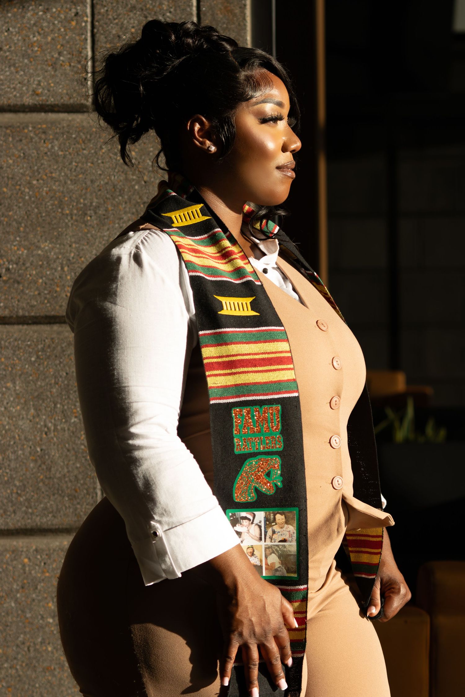A woman in a tan vest and white shirt wears a colorful Kente graduation stole in dramatic sunlight.