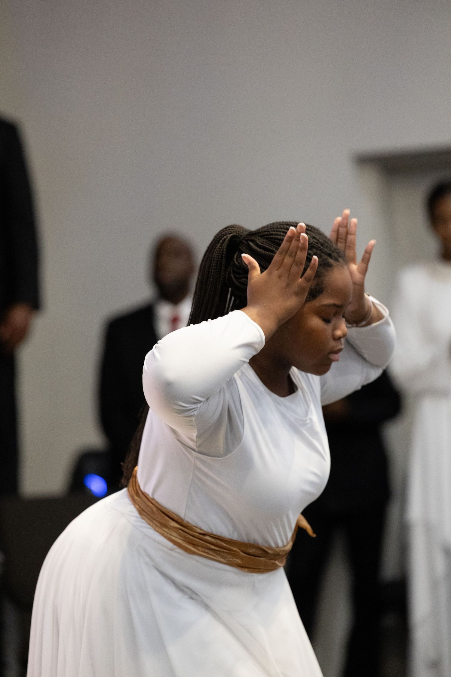 Young woman in white dress and gold sash performs liturgical dance with hands raised in prayer.