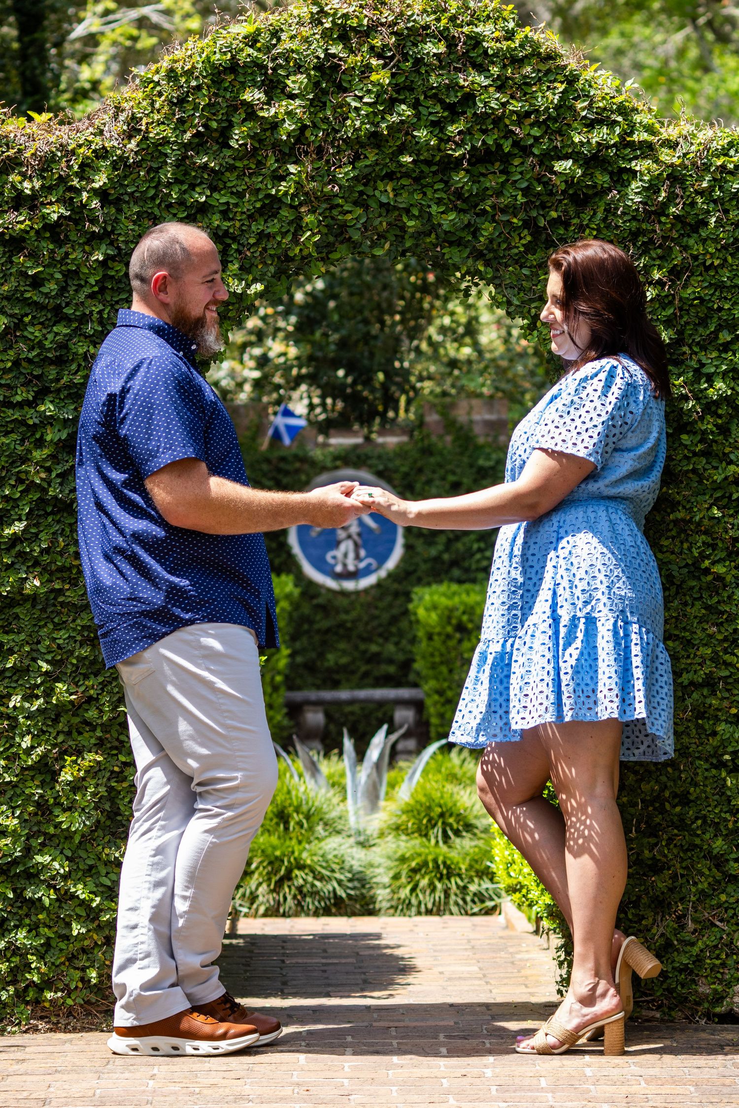 Couple holding hands in a lush garden archway, man in blue shirt and woman in blue polka dot dress.