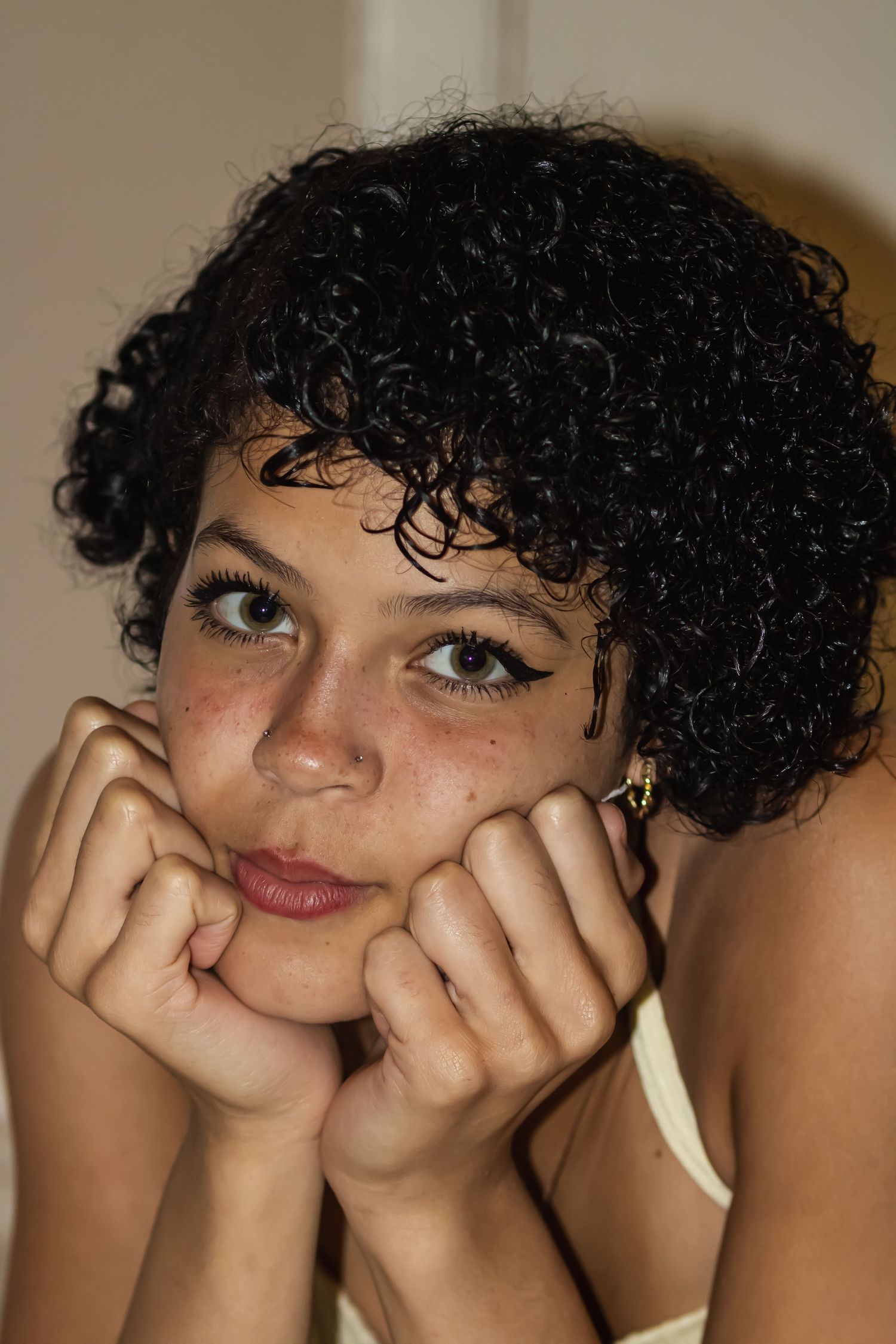 Young woman with short curly black hair resting her chin on her fists, wearing a gold earring and light top.