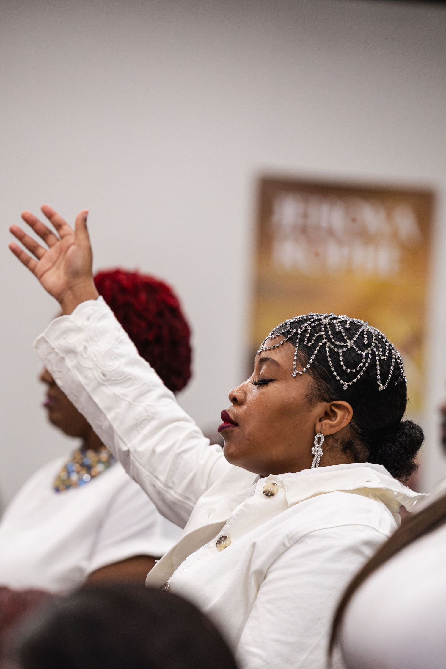 Woman in white outfit with pearl headpiece raises hand during a gospel church service.