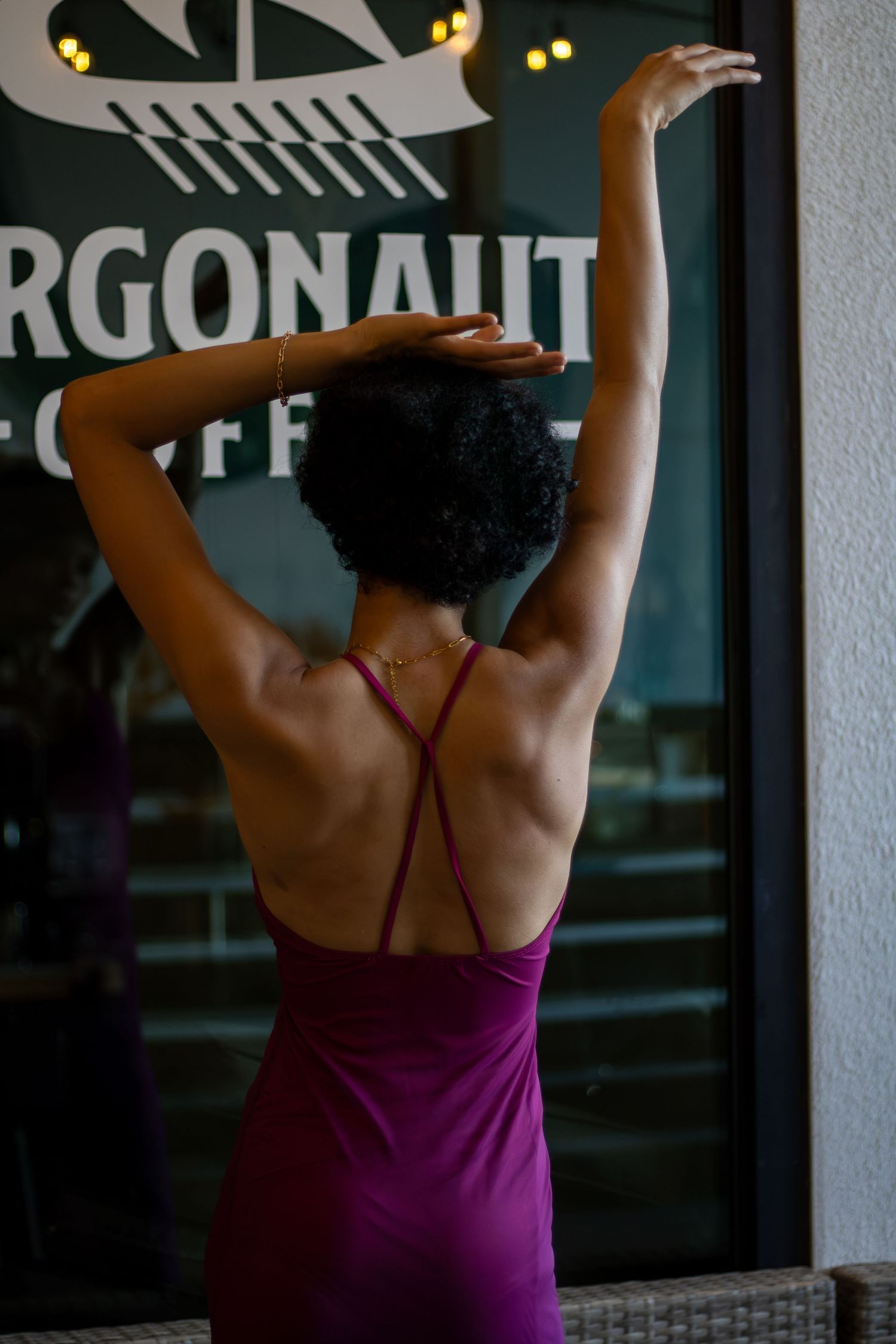 Woman in purple backless dress with arms raised, standing outside Argonaut Coffee shop.