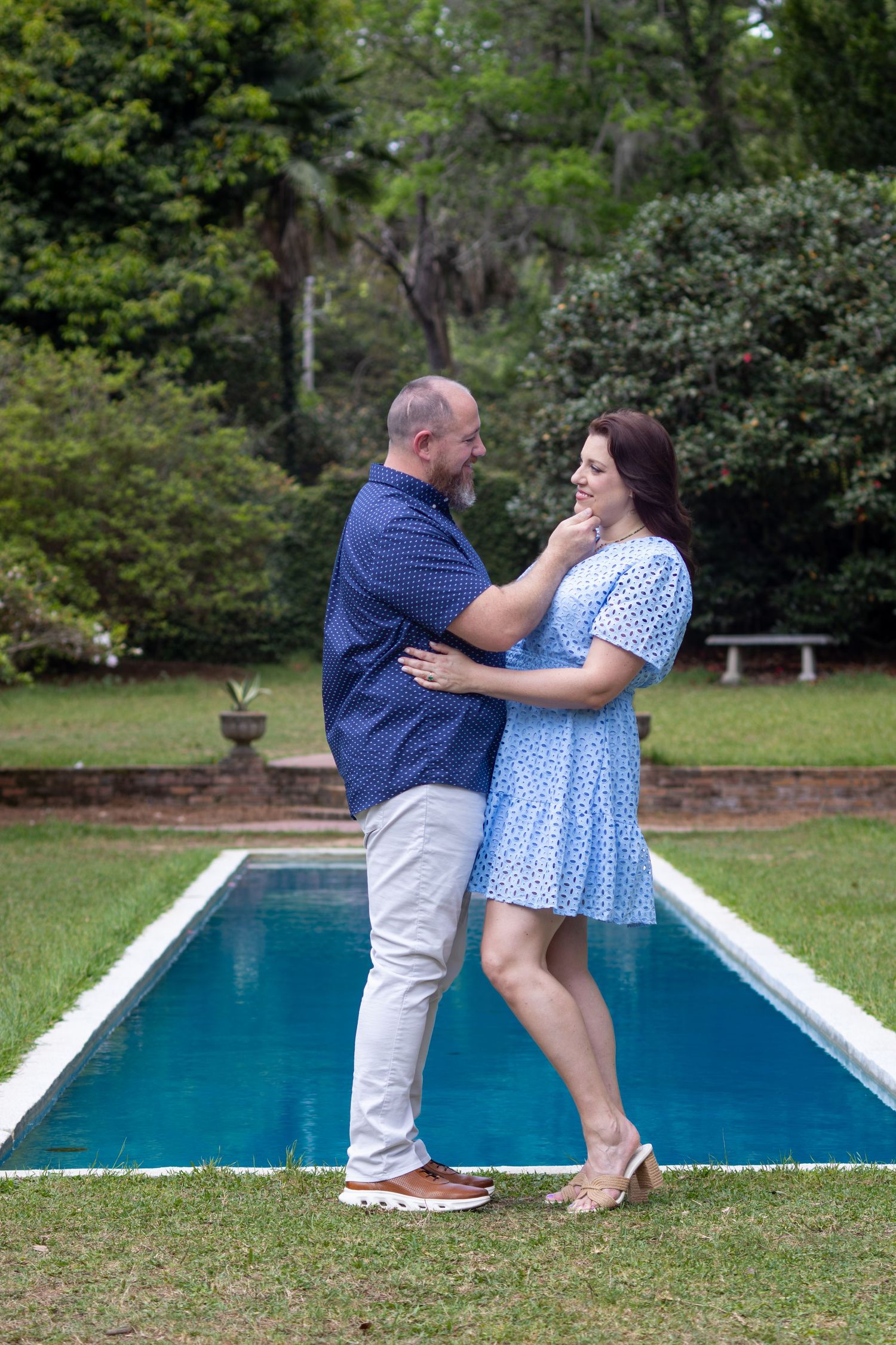 Couple embracing affectionately by a garden pool, woman in blue dress and man in navy shirt, surrounded by lush greenery.