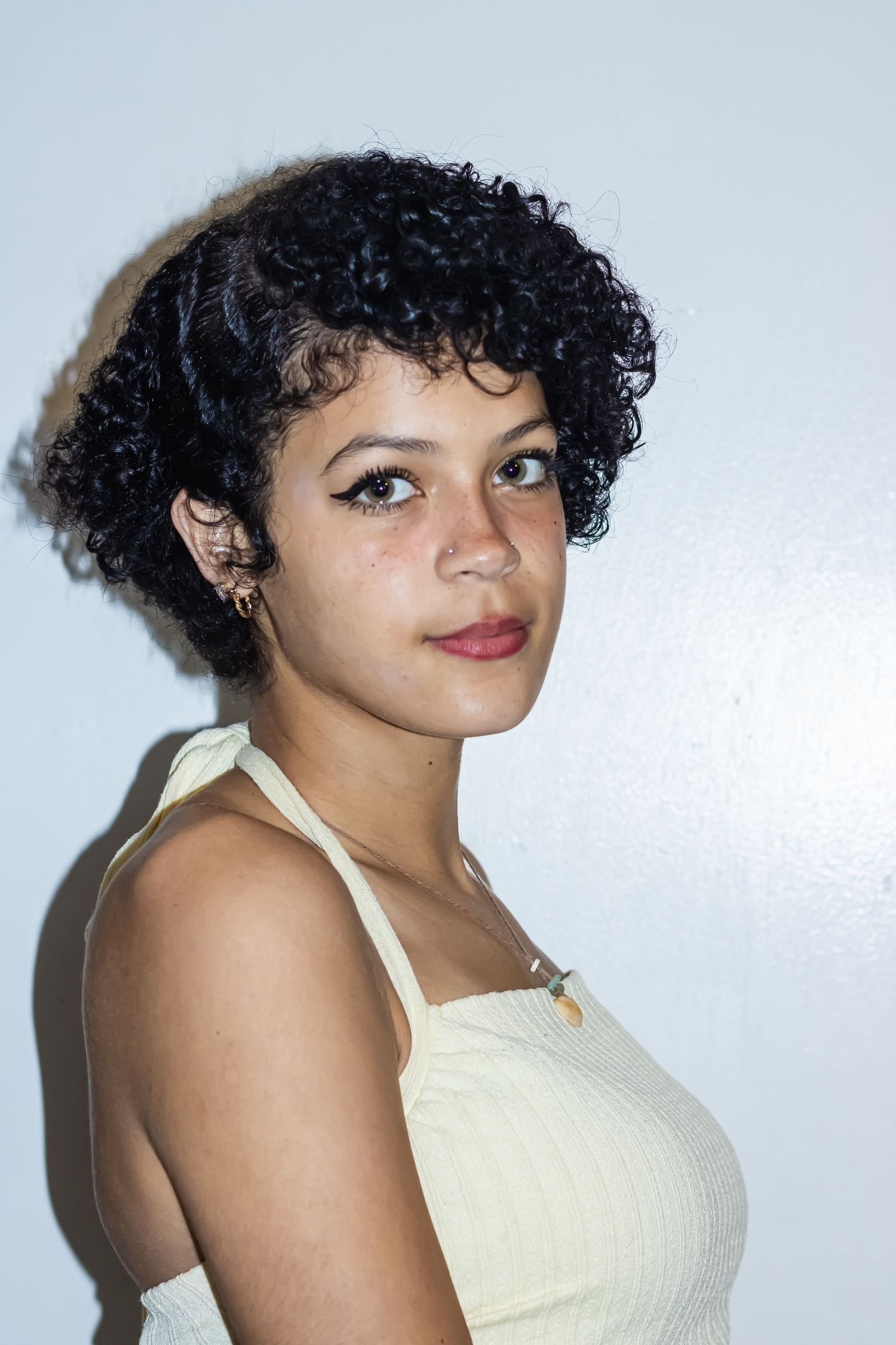 Young woman with short curly black hair wearing a cream halter top, posing against a white wall.