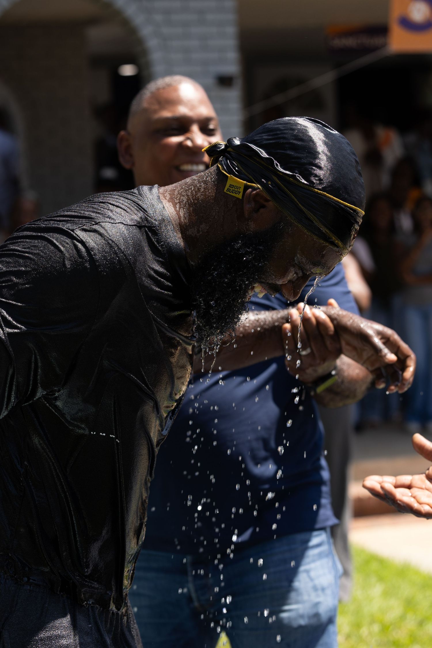 A man cools off by splashing water on his face outdoors on a sunny day while others watch nearby.