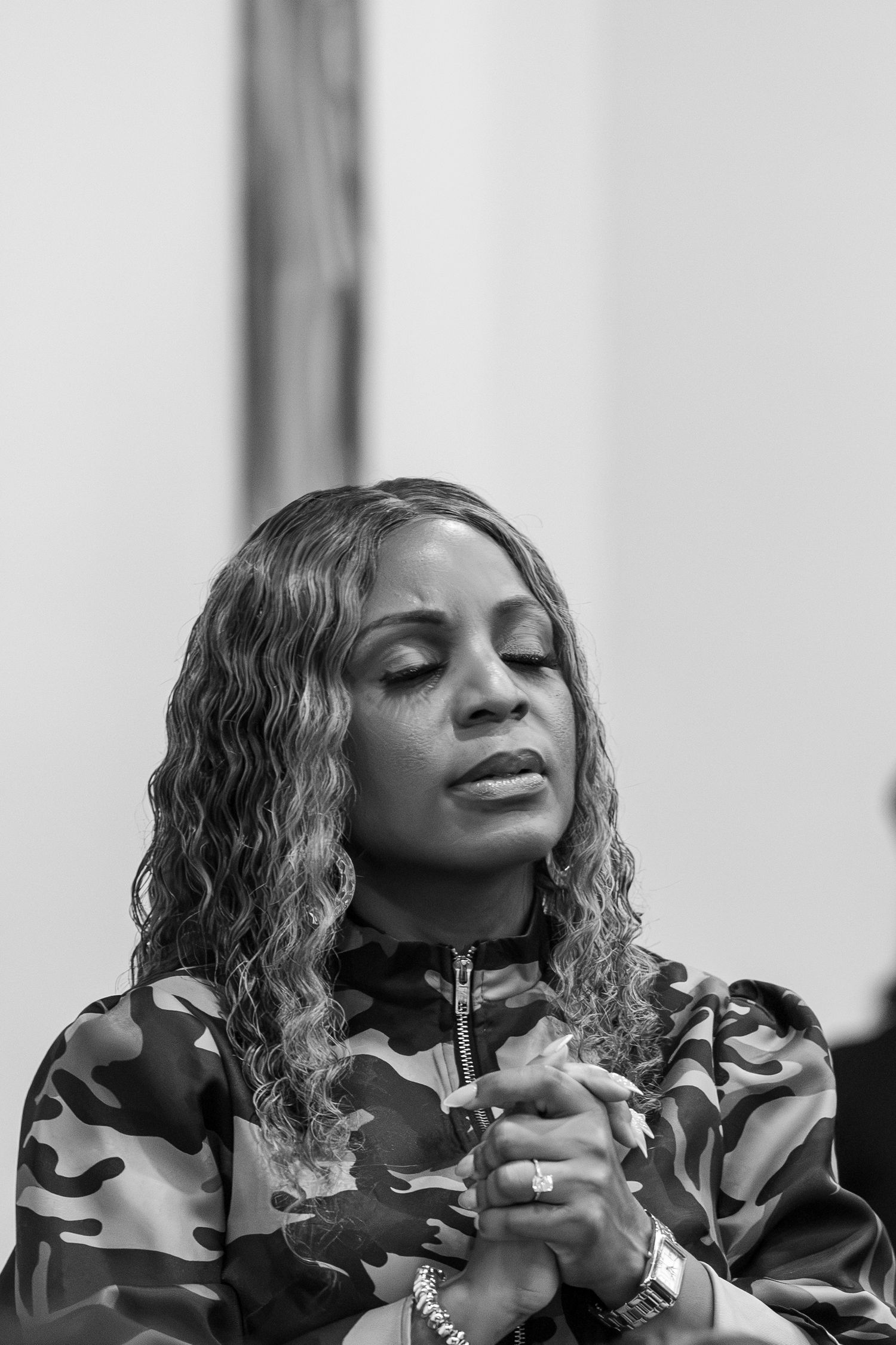 Black and white portrait of a woman with curly hair in camo jacket, eyes closed, hands clasped in prayer.